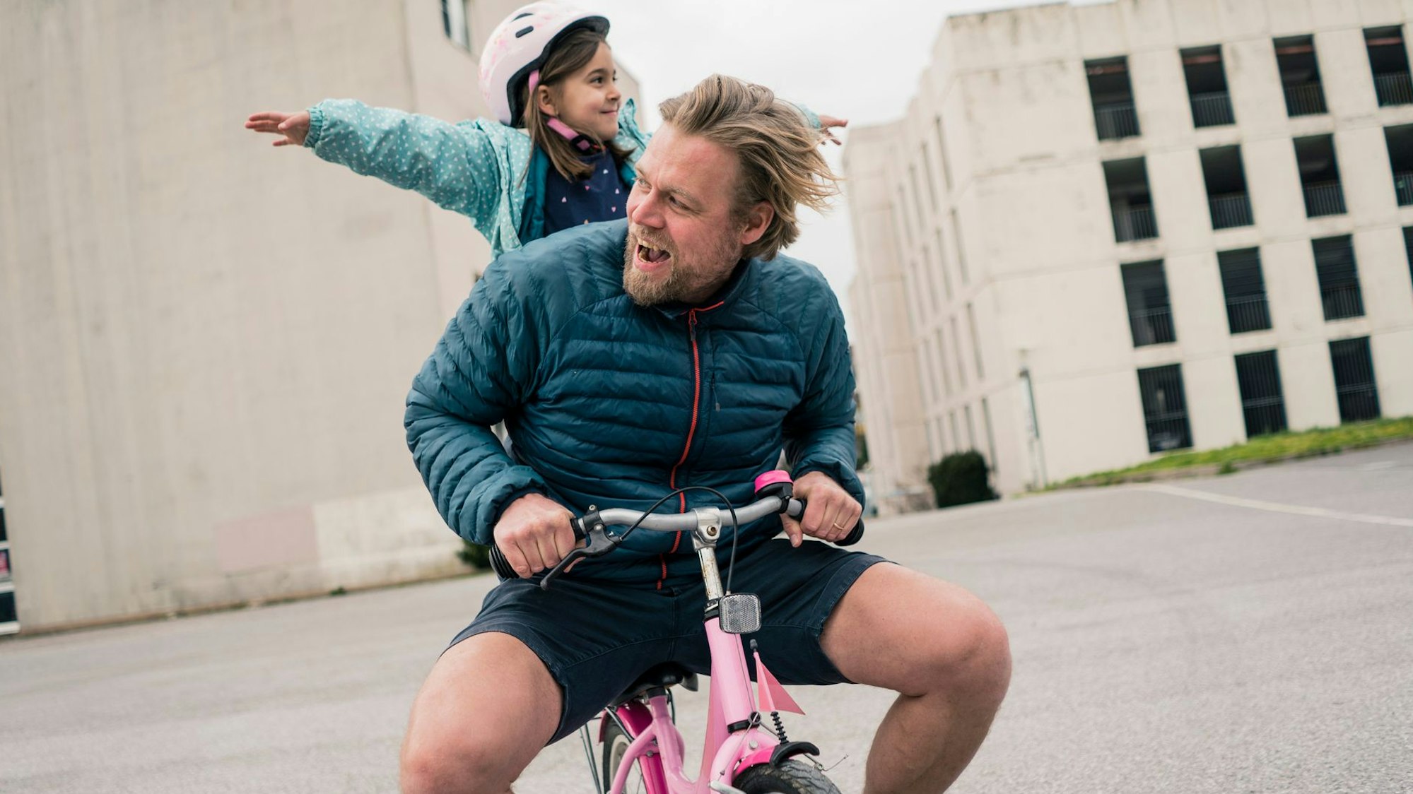 Ein junger Mann sitzt auf einem pink-farbenen Kinderrad, auf dem Gepäckträger steht ein glückliches junges Mädchen mit Helm und streckt die Arme aus.