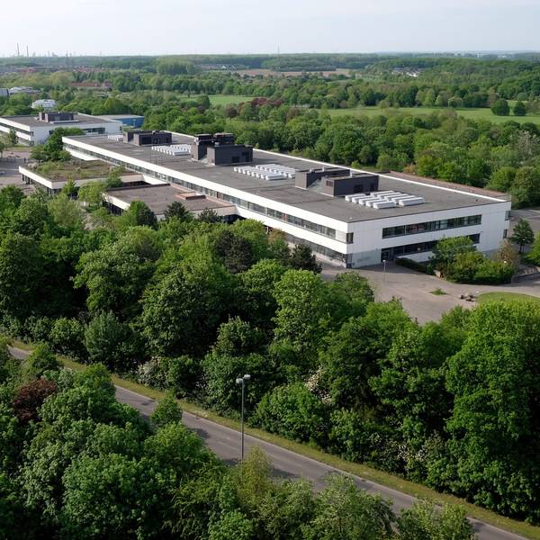Ein langgezogenes zweistöckiges Gebäude mit Flachdach ist von oben zu sehen, drumherum sind Bäume und Wiesen und eine Straße.