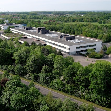 Ein langgezogenes zweistöckiges Gebäude mit Flachdach ist von oben zu sehen, drumherum sind Bäume und Wiesen und eine Straße.