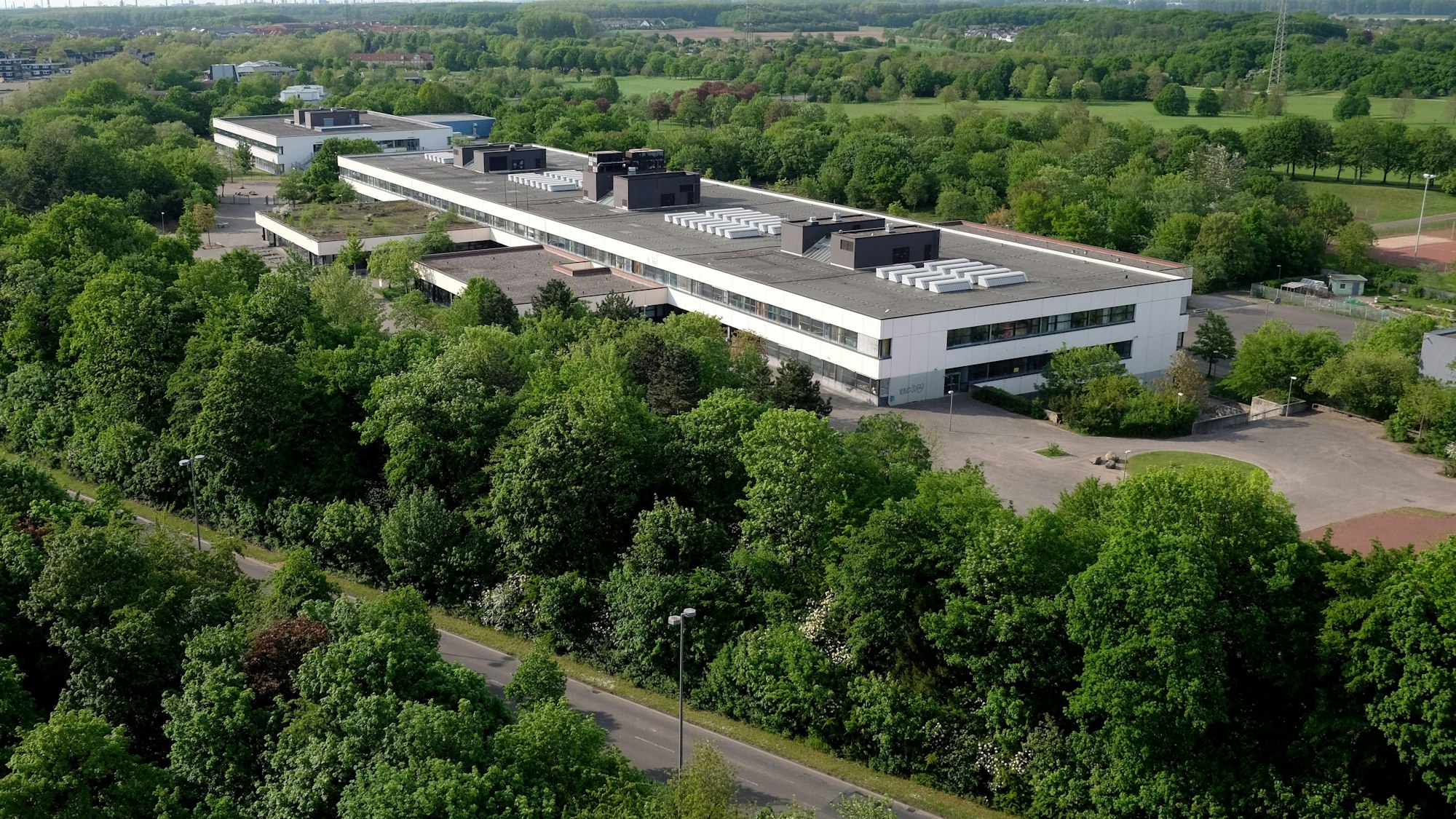 Ein langgezogenes zweistöckiges Gebäude mit Flachdach ist von oben zu sehen, drumherum sind Bäume und Wiesen und eine Straße.
