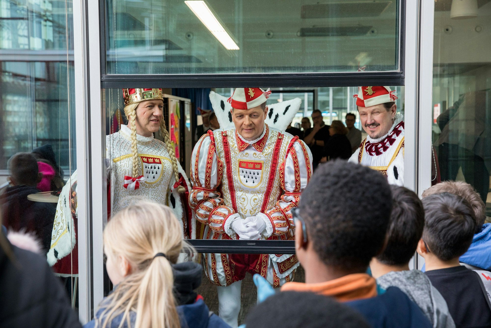 Das Kölner Dreigestirn guckt aus einem Fenster, vor dem eine Schulklasse steht.