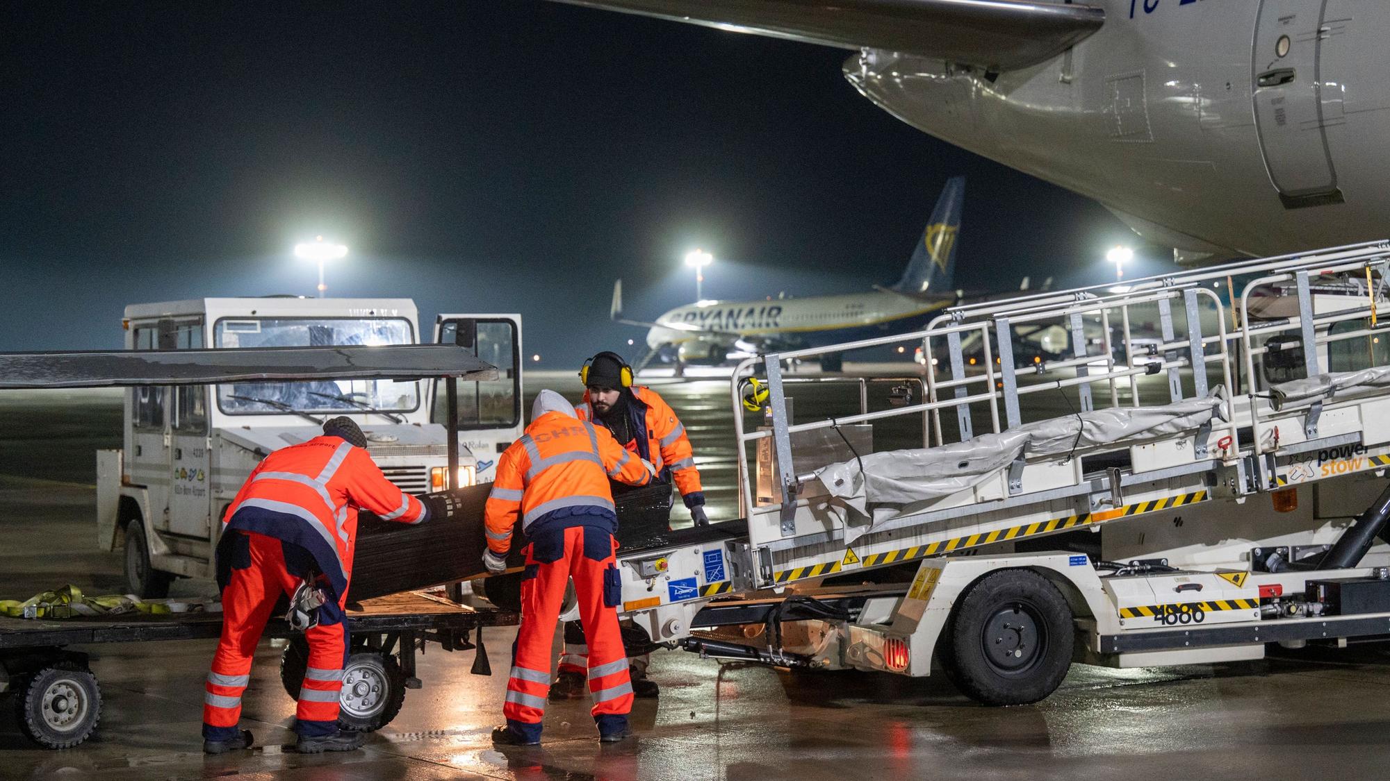 Ein Sarg wird von drei Mitarbeitern der Gepäckabfertigung auf ein Förderband verladen, das in den Innenraum eines Flugzeugs führt.