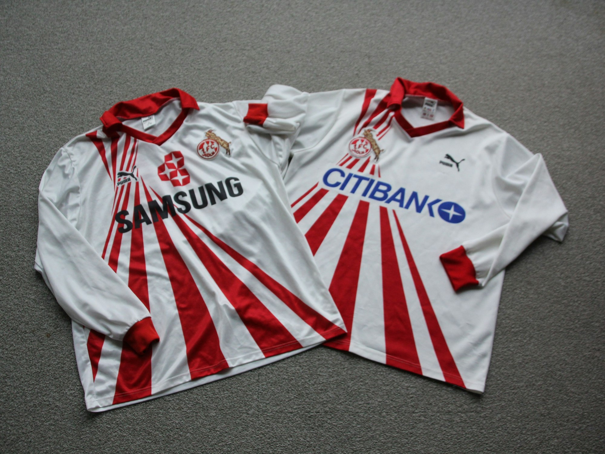 Zwei Trikots des 1. FC Köln mit dem gleichen Design, aber unterschiedlichem Sponsoren-Aufdruck: Weiß, mit roten Strahlen, die von oben nach unten verlaufen.