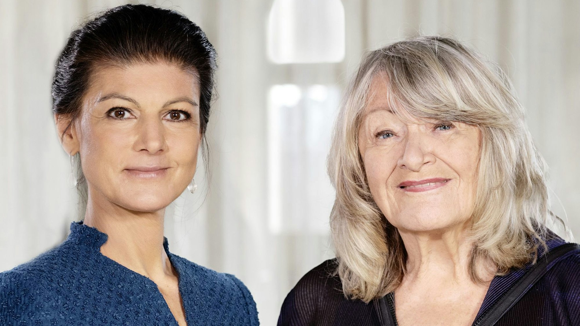 Die Politikerin Sahra Wagenknecht und die Journalistin Alice Schwarzer stehen in einem lichtdurchfluteten Innenraum nebeneinander und lächeln in die Kamera.