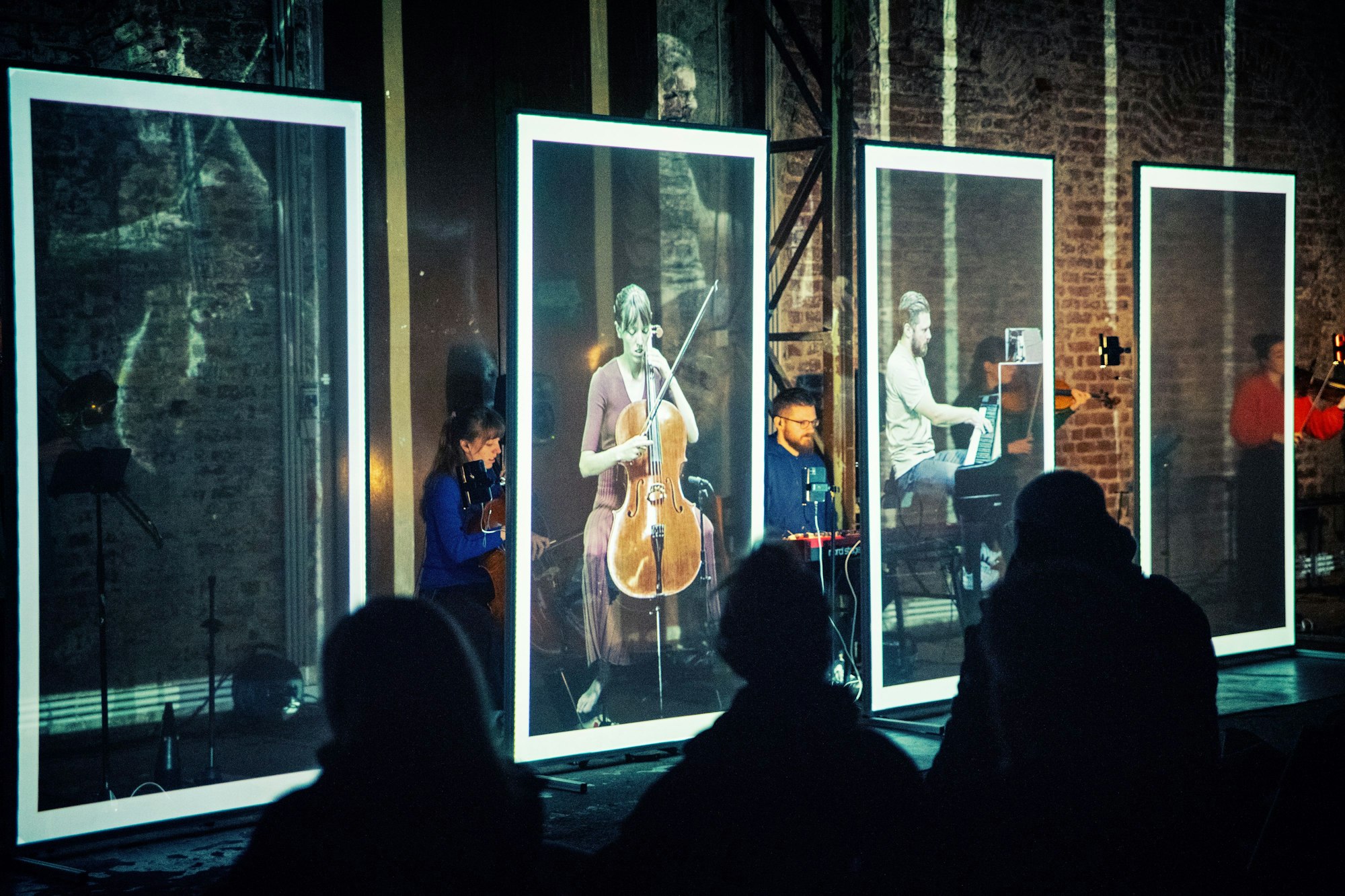 Das Bild zeigt. wie die Musiker des Ensembles Electronic ID hinter ihren Projektionen verschwinden. Vor ihnen sind in einer Reihe transparente Kästen in Fotoform, in denen Videos von ihnen beim Spielen ihrer Instrumente gezeigt werden