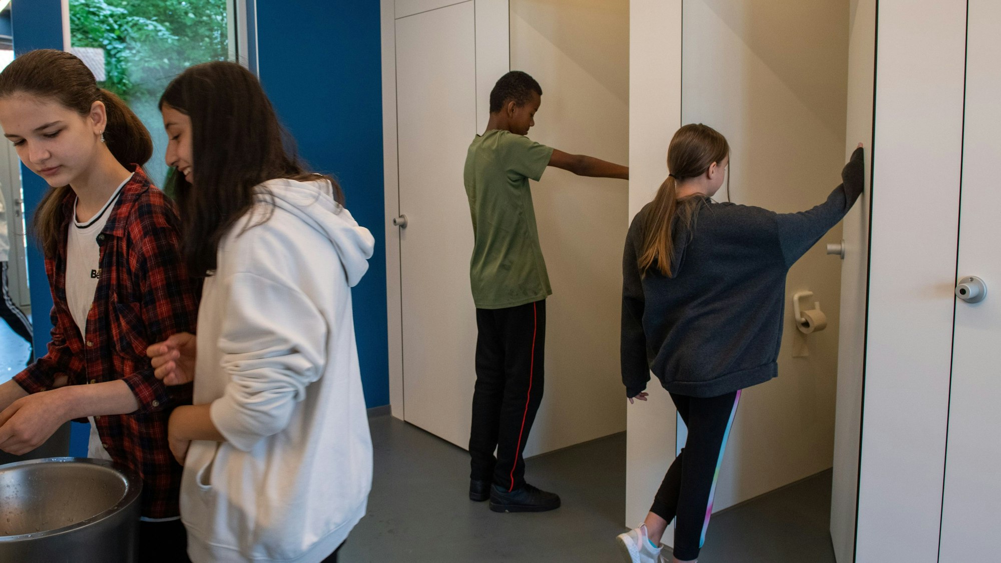 Ein Junge und ein Mädchen gehen durch eine Tür auf die Toilette. Im Vordergrund waschen sich zwei Mädchen die Hände.