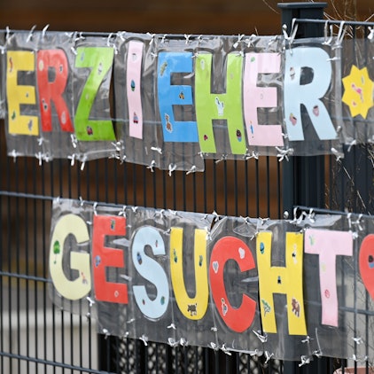 Mit bunten Buchstaben sind die Worte Erzieher*in gesucht· geformt, die an einem Zaun einer Kindertagesstätte befestigt sind.