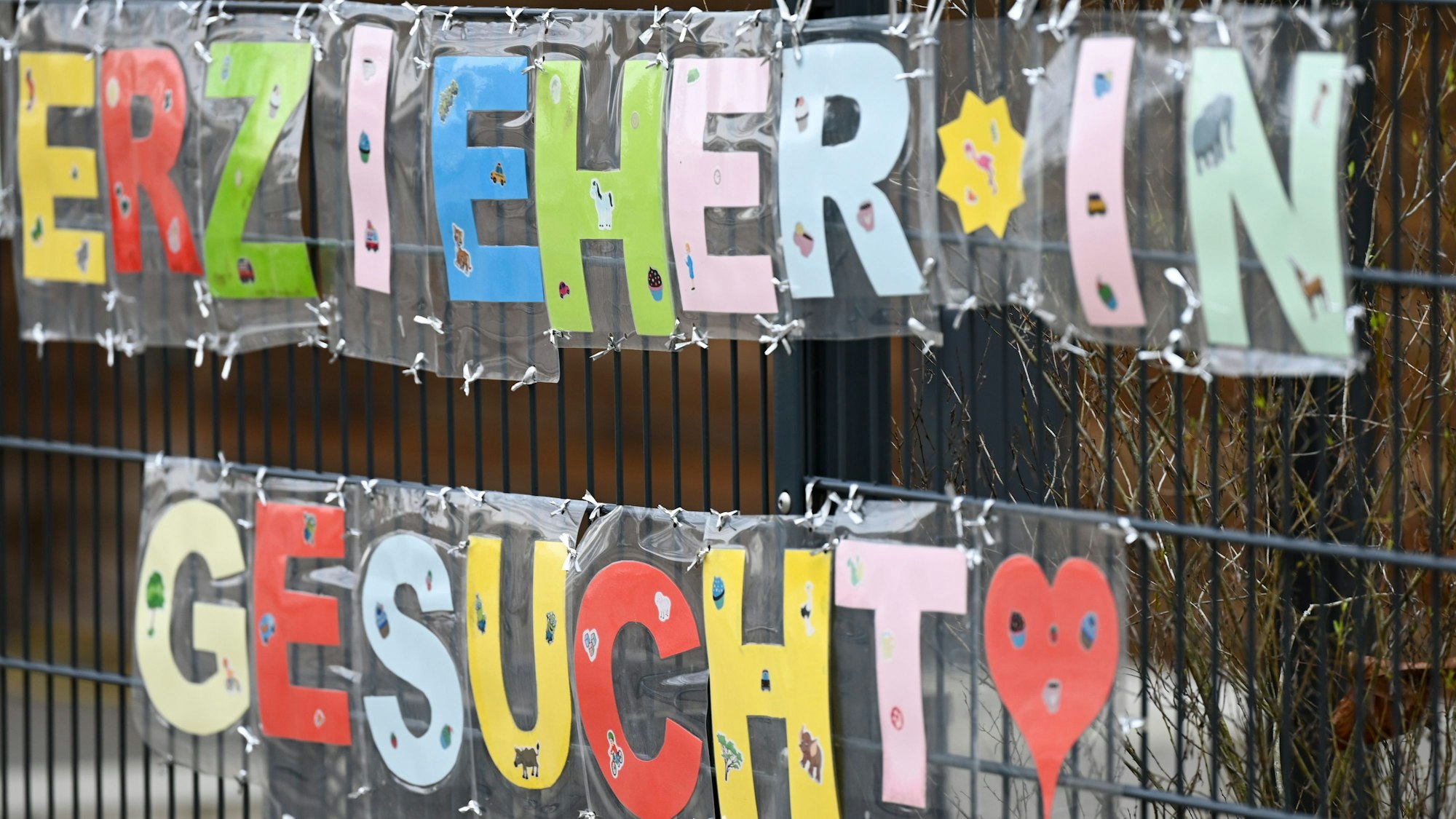 Mit bunten Buchstaben sind die Worte „Erzieher*in gesucht“ geformt, die an einem Zaun einer Kindertagesstätte hängen.