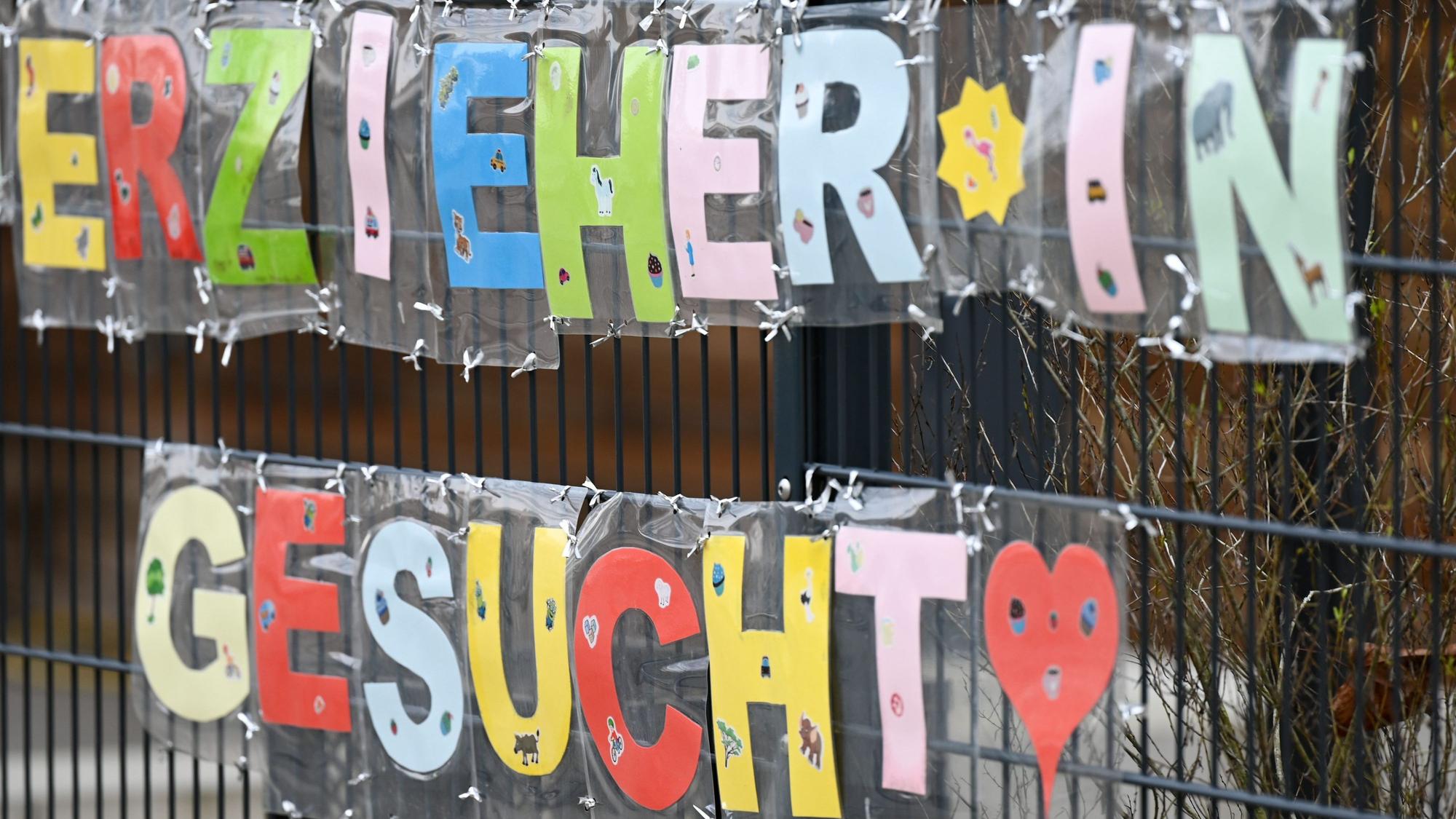 Mit bunten Buchstaben sind die Worte Erzieher*in gesucht· geformt, die an einem Zaun einer Kindertagesstätte befestigt sind.