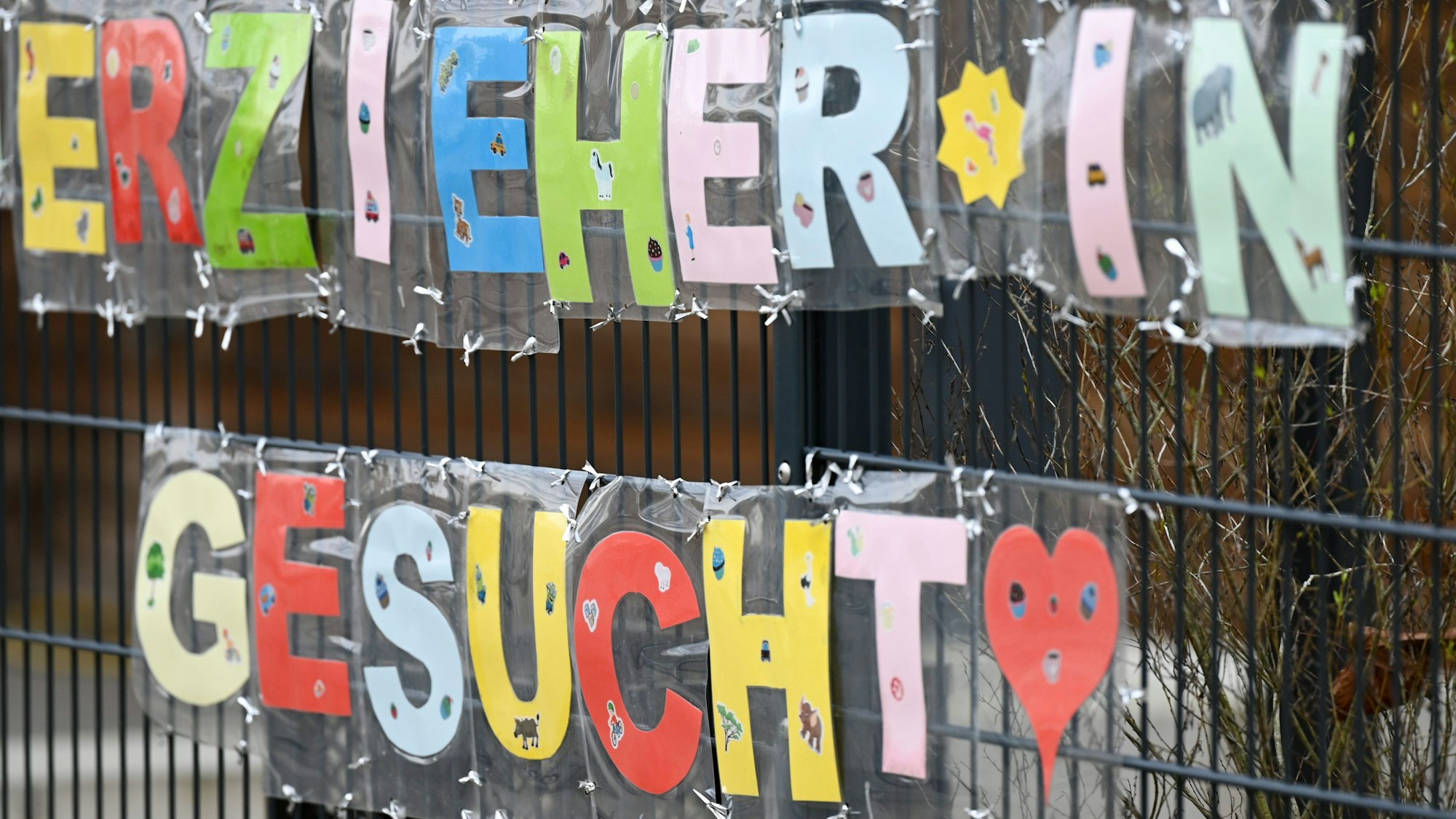 Mit bunten Buchstaben sind die Worte "Erzieher*in gesucht" geschrieben, die an einem Zaun einer Kindertagesstätte befestigt sind.