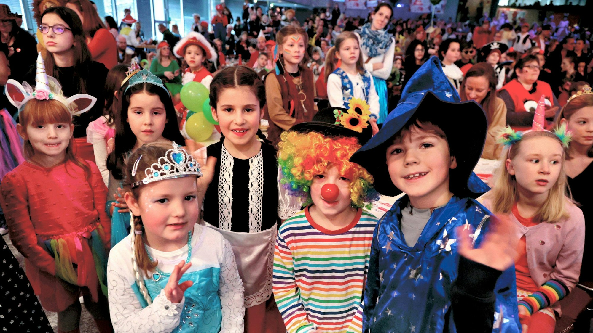 RoteSechs Kinder in Kostümen - Clowns, Prinzessinen, Einhörner und Zauberer - stehen im vollbesetzten Saal vor der Kamera und lächeln.