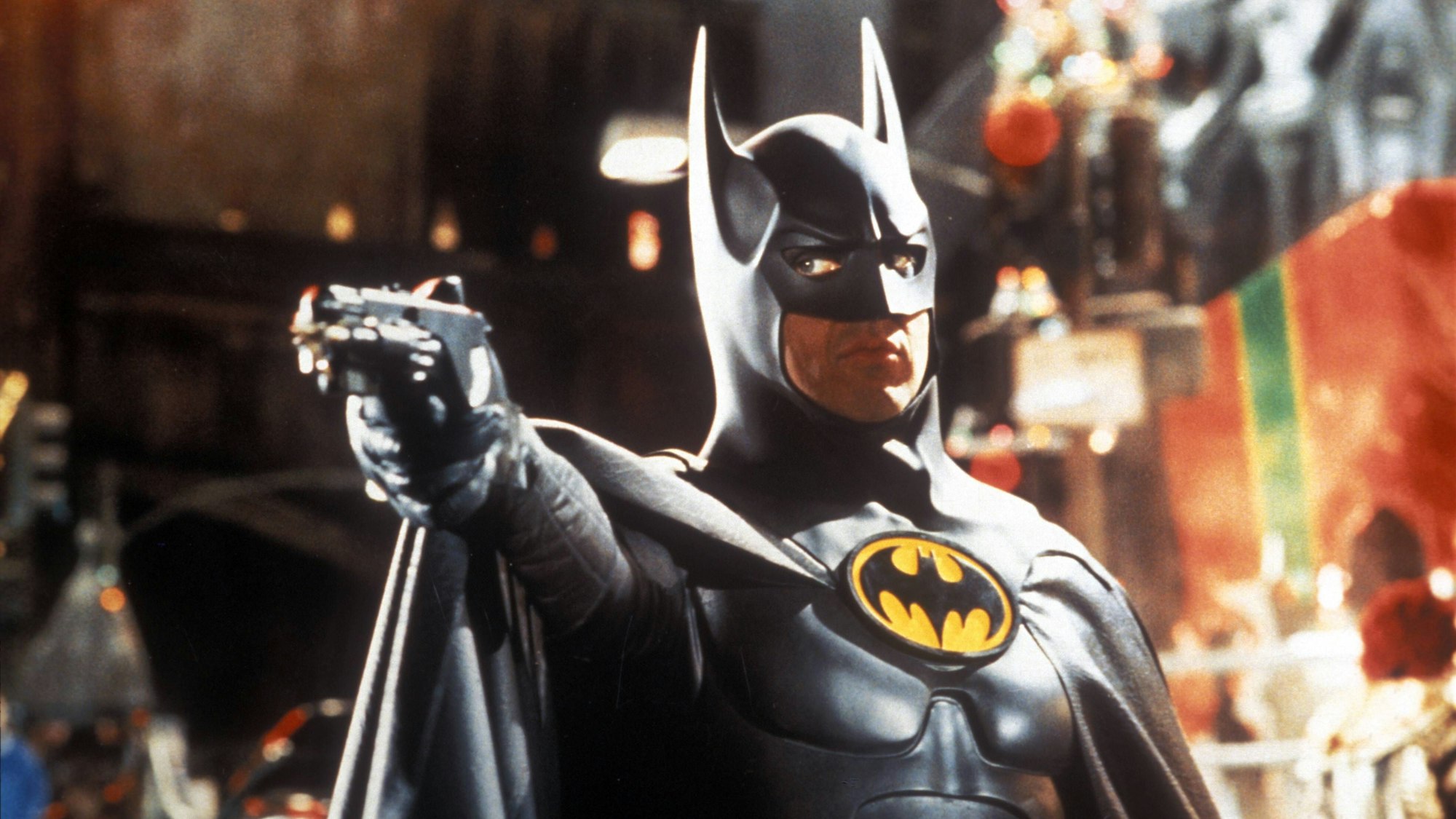 Michael Keaton als Batman, er trägt den schwarzen Batman-Anzug und hält eine Pistole in der ausgestreckten Hand.