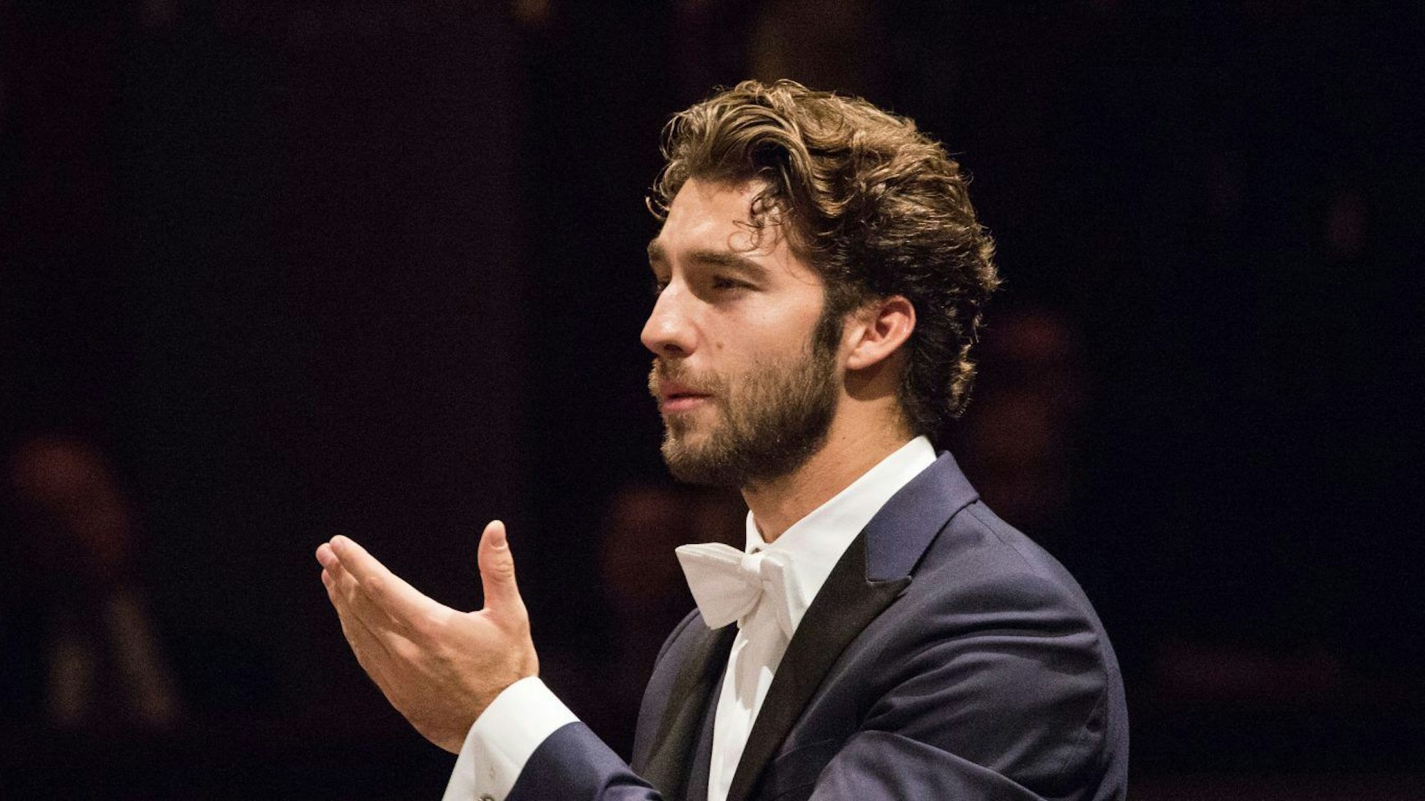 Lorenzo Viotti hat die and zum Dirigat erhoben. Er trägt einen Anzug mit weißer Fliege.