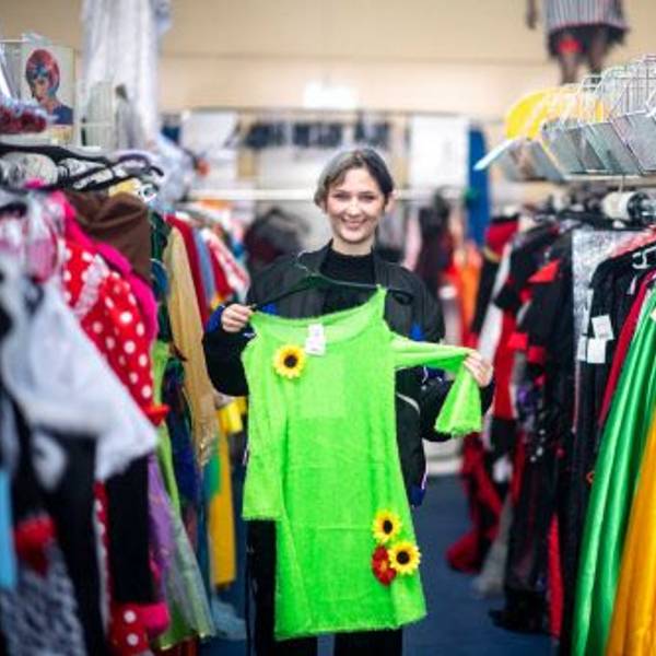 Elena Pintus hält ein grünes Kleid mit Sonnenblumen hoch. Sie steht in einem Gang zwischen Kleiderstangen mit Karnevalskostümen.