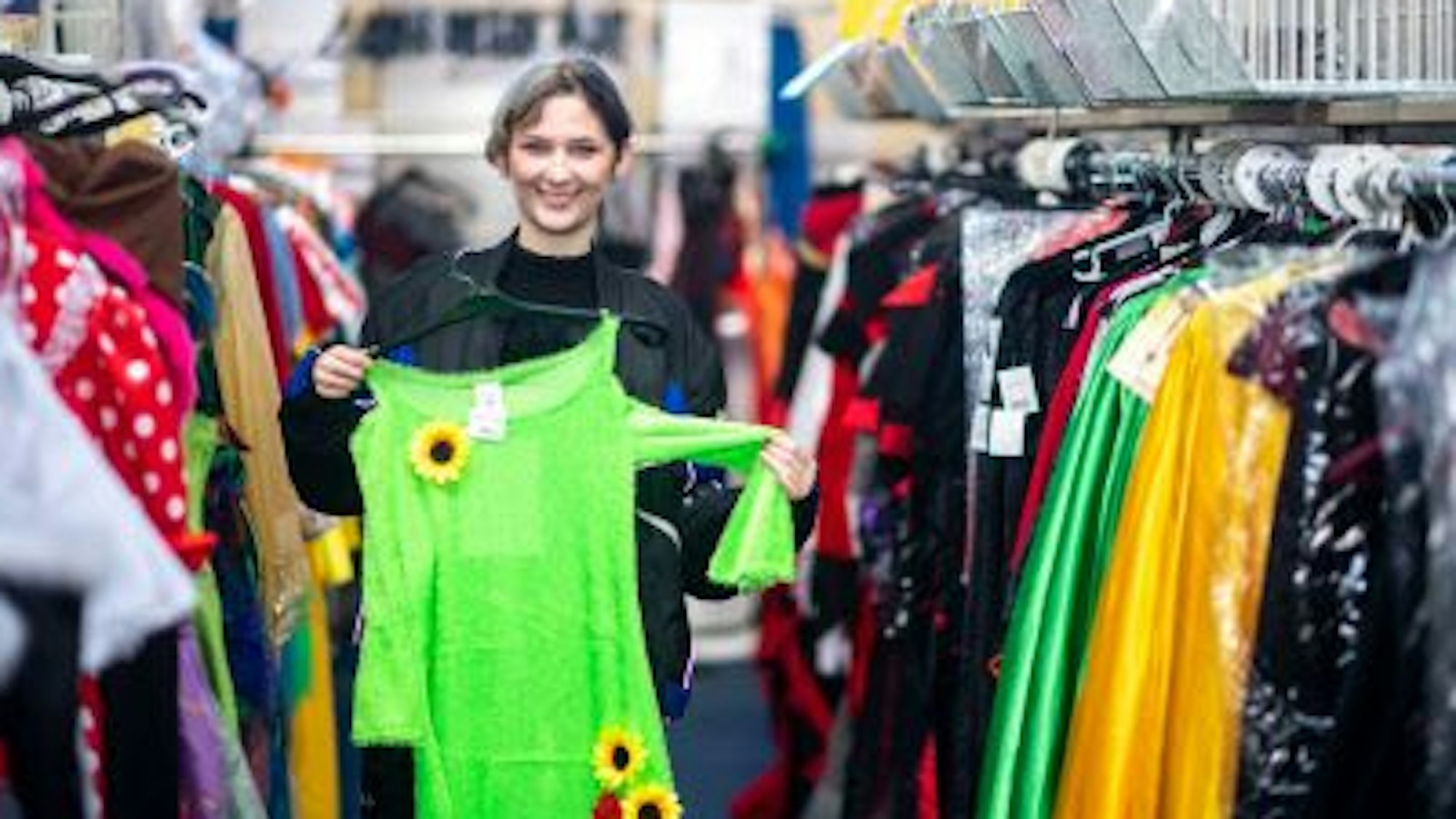 Elena Pintus hält ein grünes Kleid mit Sonnenblumen hoch. Sie steht in einem Gang zwischen Kleiderstangen mit Karnevalskostümen.