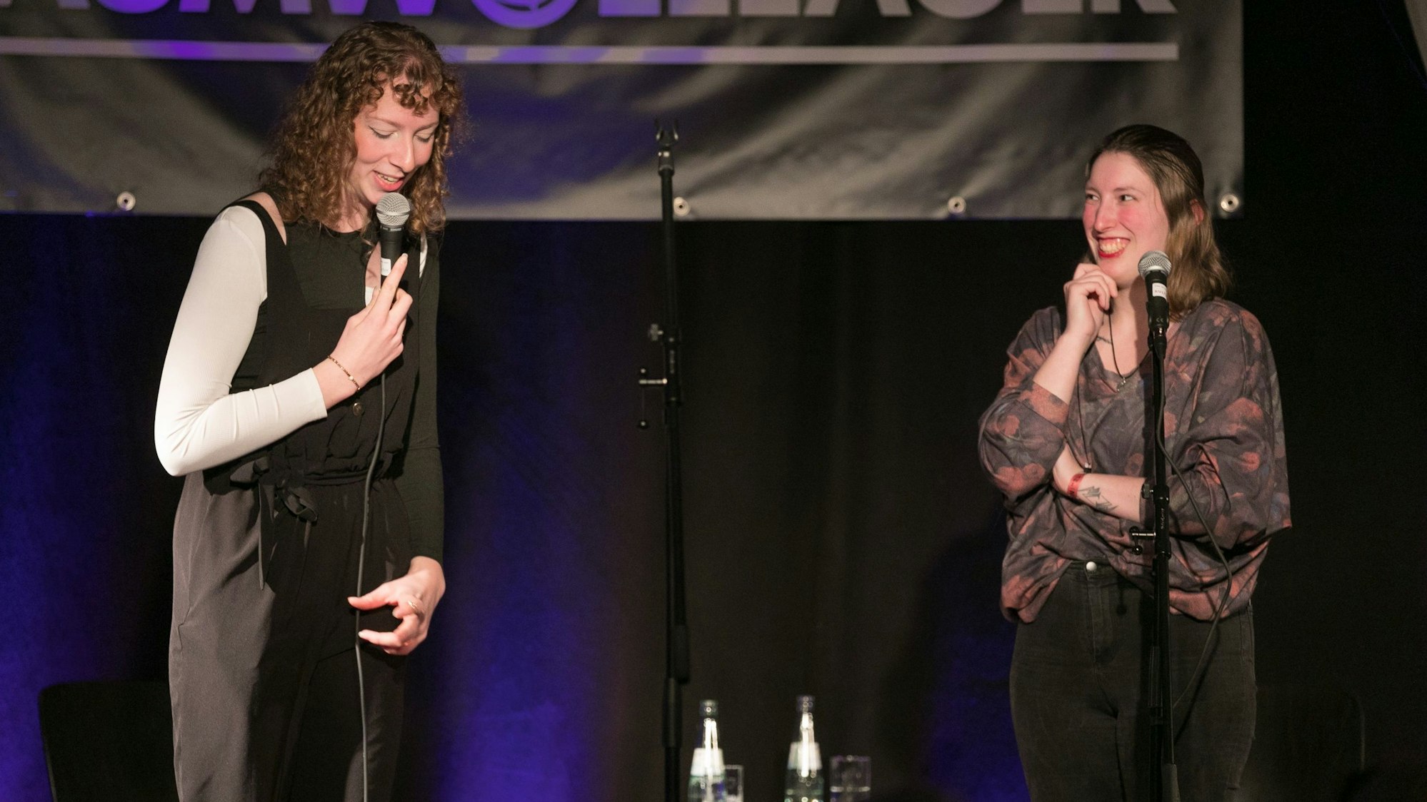 Zwei Frauen stehen auf einer Bühne: Eine spricht in ein Mikrofon, die andere schaut sie an und lacht.