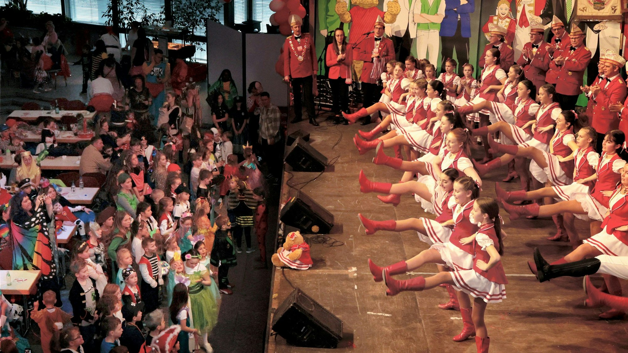 Rechts auf der Bühne tanzen drei Dutzend junge Tänzerinnen in Rot und Weiß, vor der Bühne stehen doppelt so viele verkleidete Kinder und schauen ihnen zu.