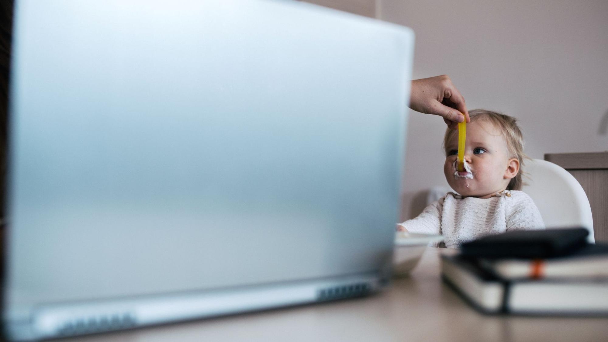 Eine Hand kommt hinter dem Laptop hervor und füttert ein Baby.