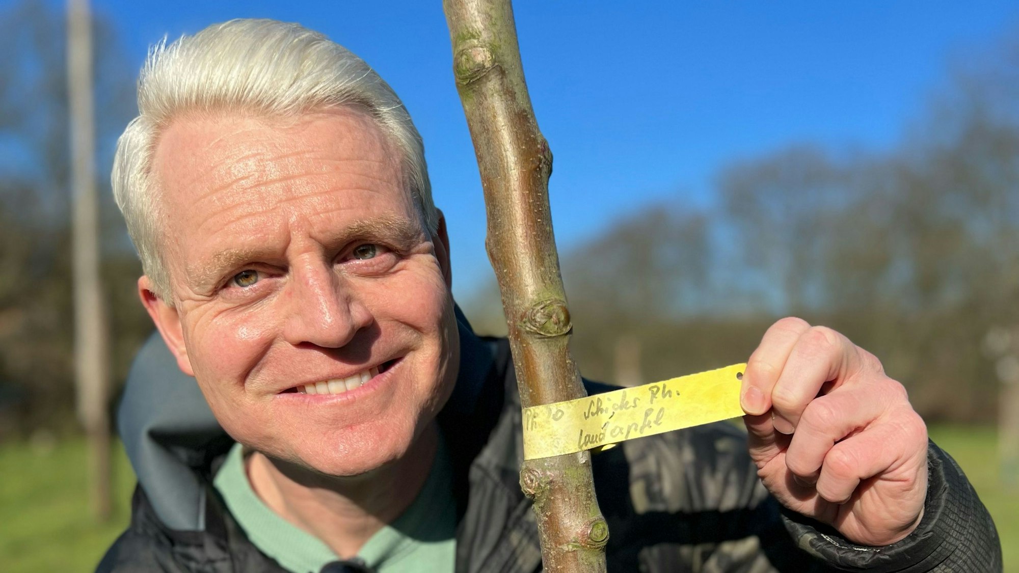 Guido Cantz lächelt in die Kamera und zeigt die Beschriftung des gepflanzten Apfelbaums.