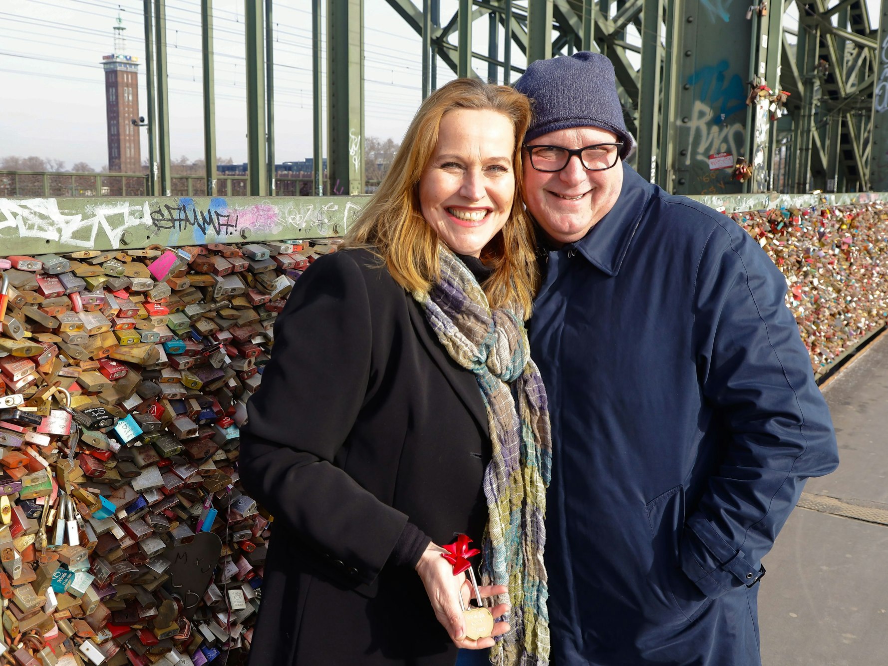 Nicole Sterck-Schultz und Frank Schultz stehen auf der Hohenzollernbrücke vor den Liebesschlössern und halten ihr Schloss in der Hand.