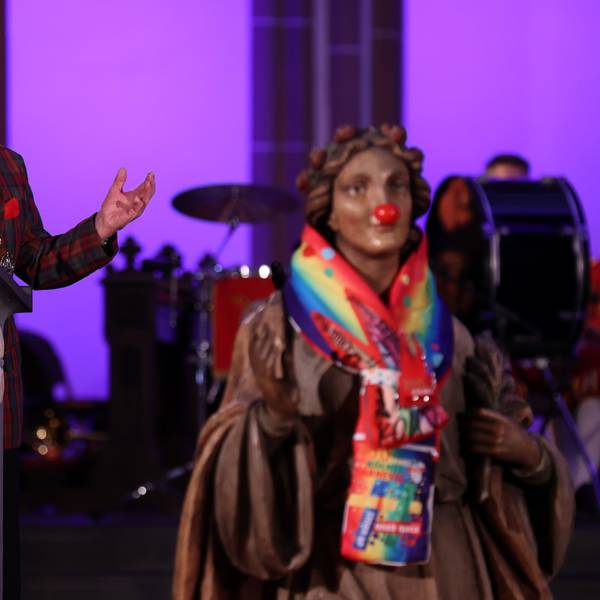 Ludwig Sebus steht am Rednerpult und hält die Predigt, im Vordergrund ist die Skulptur der heiligen Agnes mit Clownsnase zu sehen.