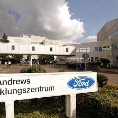 Abgebildet ist ein großes, U-förmiges Gebäude. Im Vordergrund steht ein Schild mit der Aufschrift „John Andrews Entwicklungszentrum“ mit dem blauen, ovalen Ford-Logo.