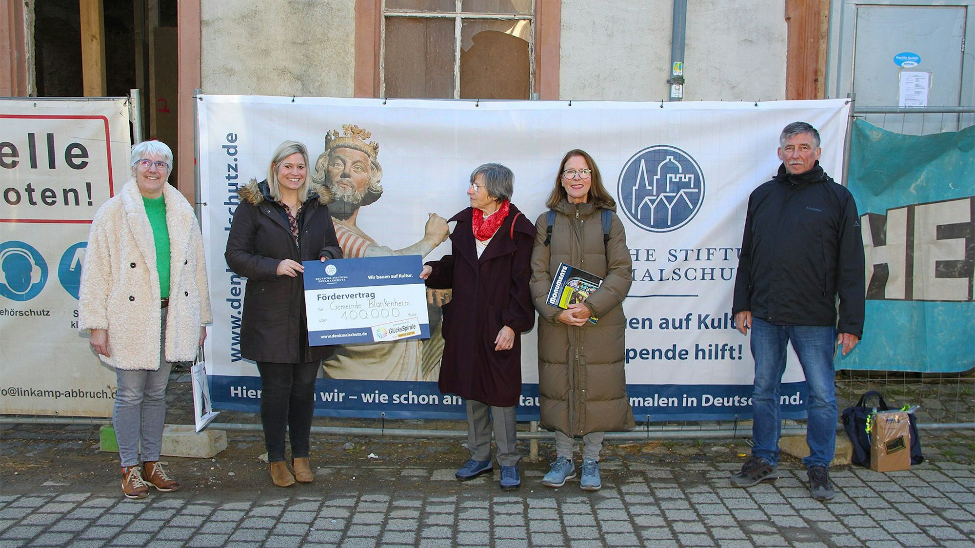 Fünf Personen stehen vor einem Haus, an dessen Fassade Plakate hängen.