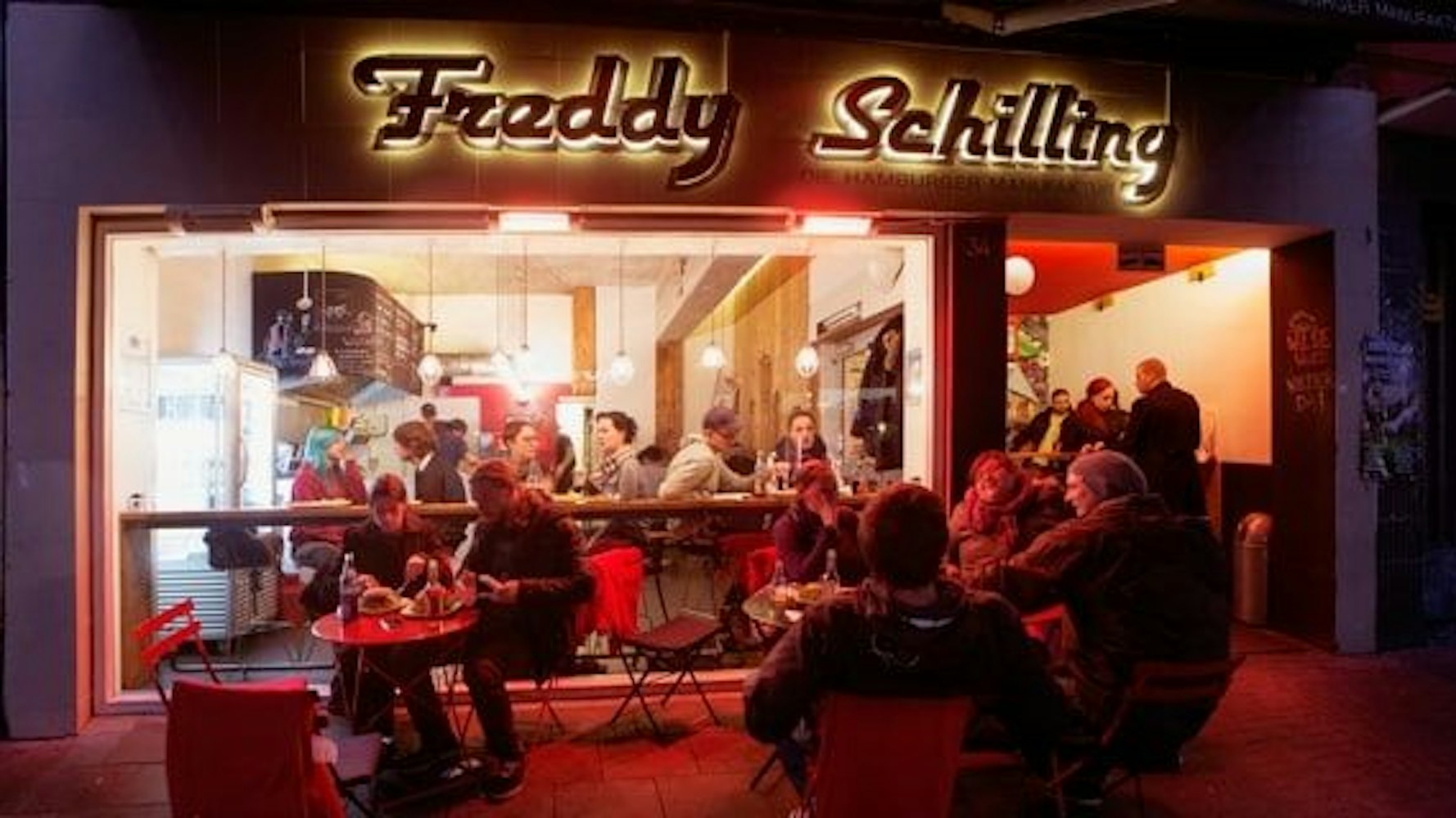 Außenansicht bei Nacht des Freddy Schilling. Menschen sitzen vor dem Laden und Essen. In Leuchtschrift ist über dem Restaurant der Name des Laden zu lesen.