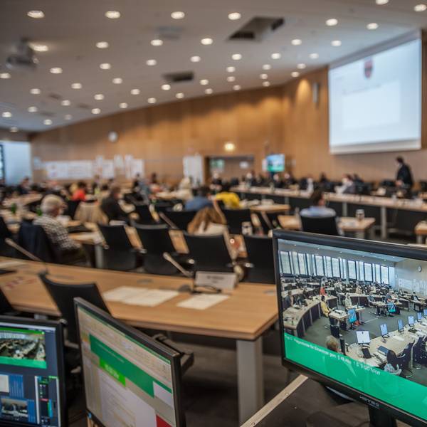 Blick in den Ratssaal, im Vordergrund ein Monitor, der das Bild der Live-Übertragung zeigt.