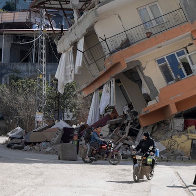 Zwei Männer in Warnwesten gehen an einem zerstörten Haus in Kirikhan vorbei.