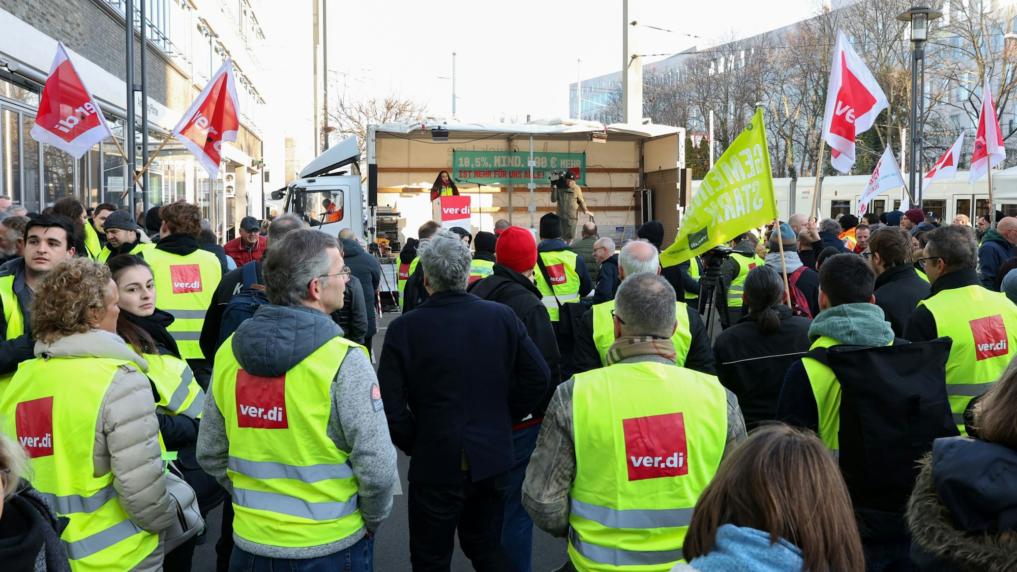 Mitarbeiter und Mitarbeiterinnen der KVB in Warnwesten stehen vor einer Bühne und haben teilweise Fahnen in der Hand.