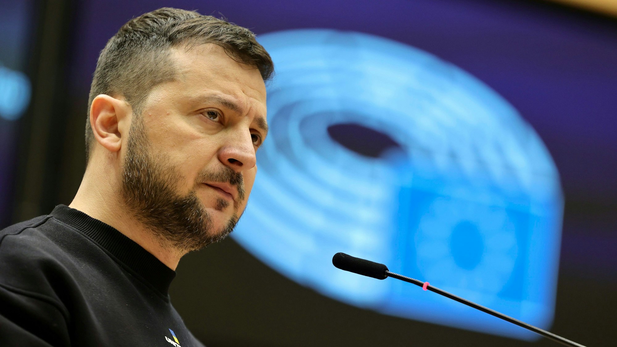 Wolodymyr Selenskyj, Präsident der Ukraine, spricht bei einem EU-Gipfel im Europäischen Rat in Brüssel. Selenskyjs ist im Profil zu sehen, vor ihm ein Mikrofon, hinter ihm eine blau ausgeleuchtete Wand.