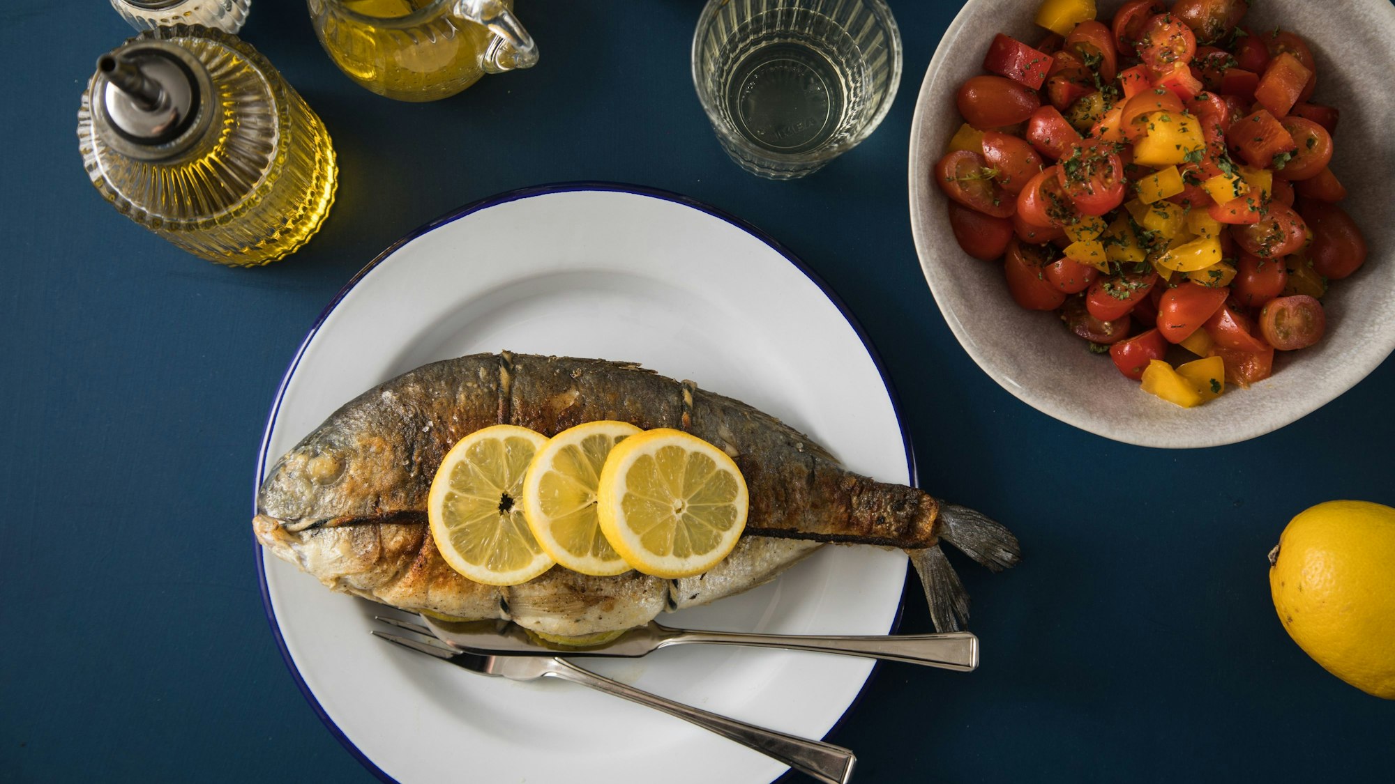 Auf einem Teller liegt eine mit Zitronenscheiben garnierte Dorade, daneben eine Schüssel mit Gemüse.