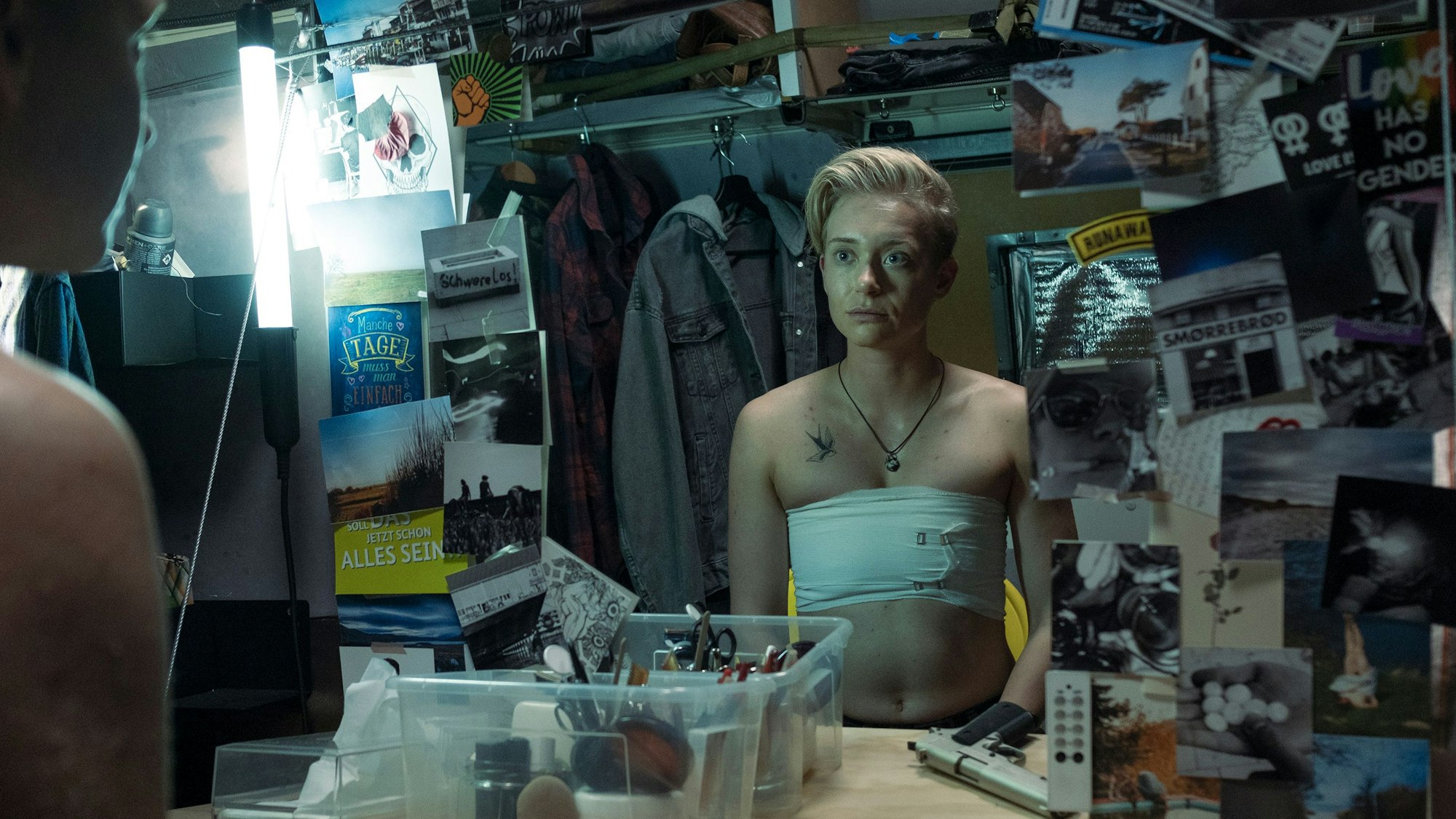 Der blonde Jonathan Perleth hat sich die Brust mit Bandagen abgebunden, sitzt ansonsten oberkörperfrei vor dem Spiegel. Um den Spiegel herum sind viele Postkarten und Fotos.