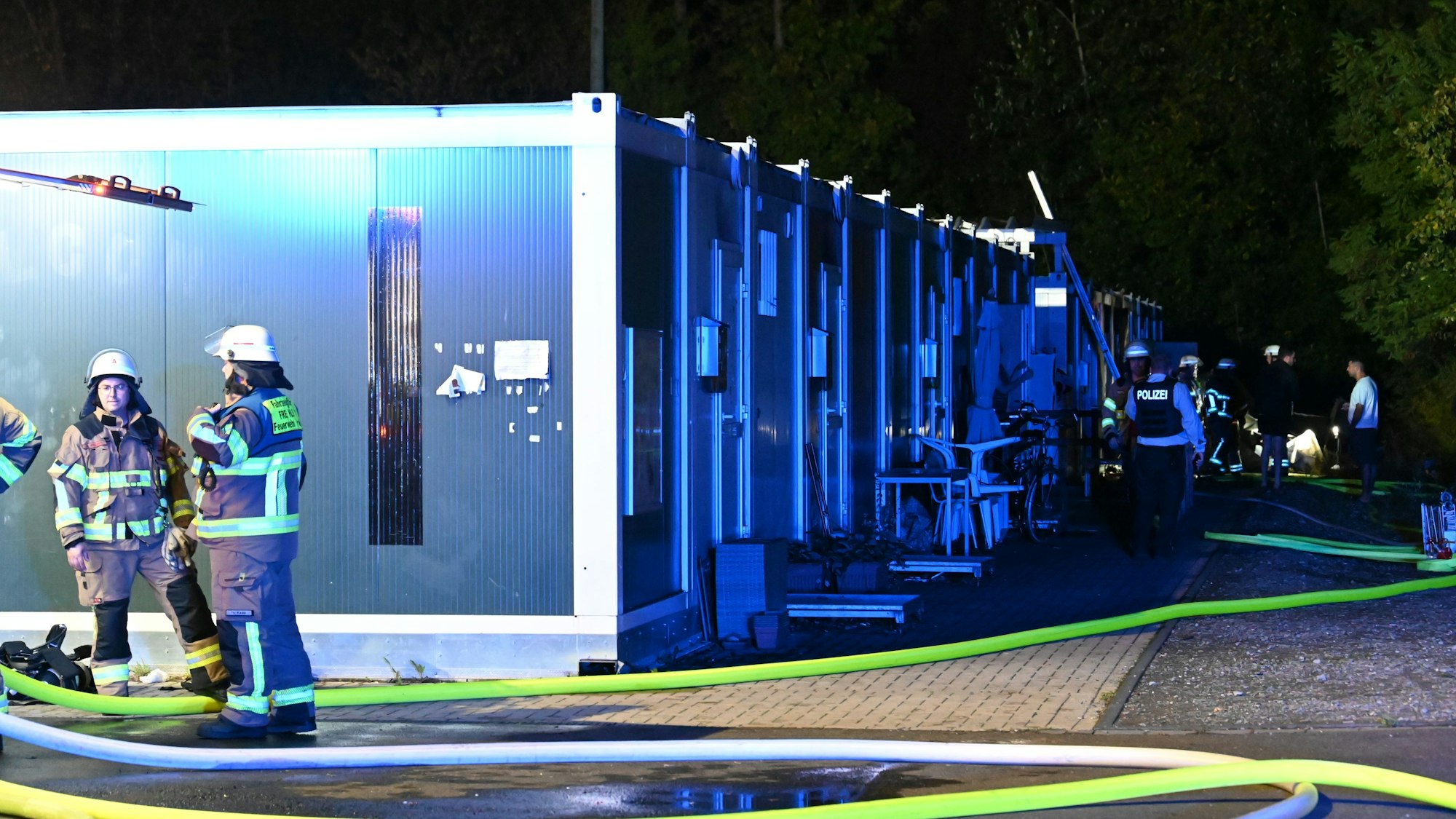 Das Foto zeigt Feuerwehrleute, die vor den Wohncontainern stehen. Der Einsatzort ist hell erleuchtet.