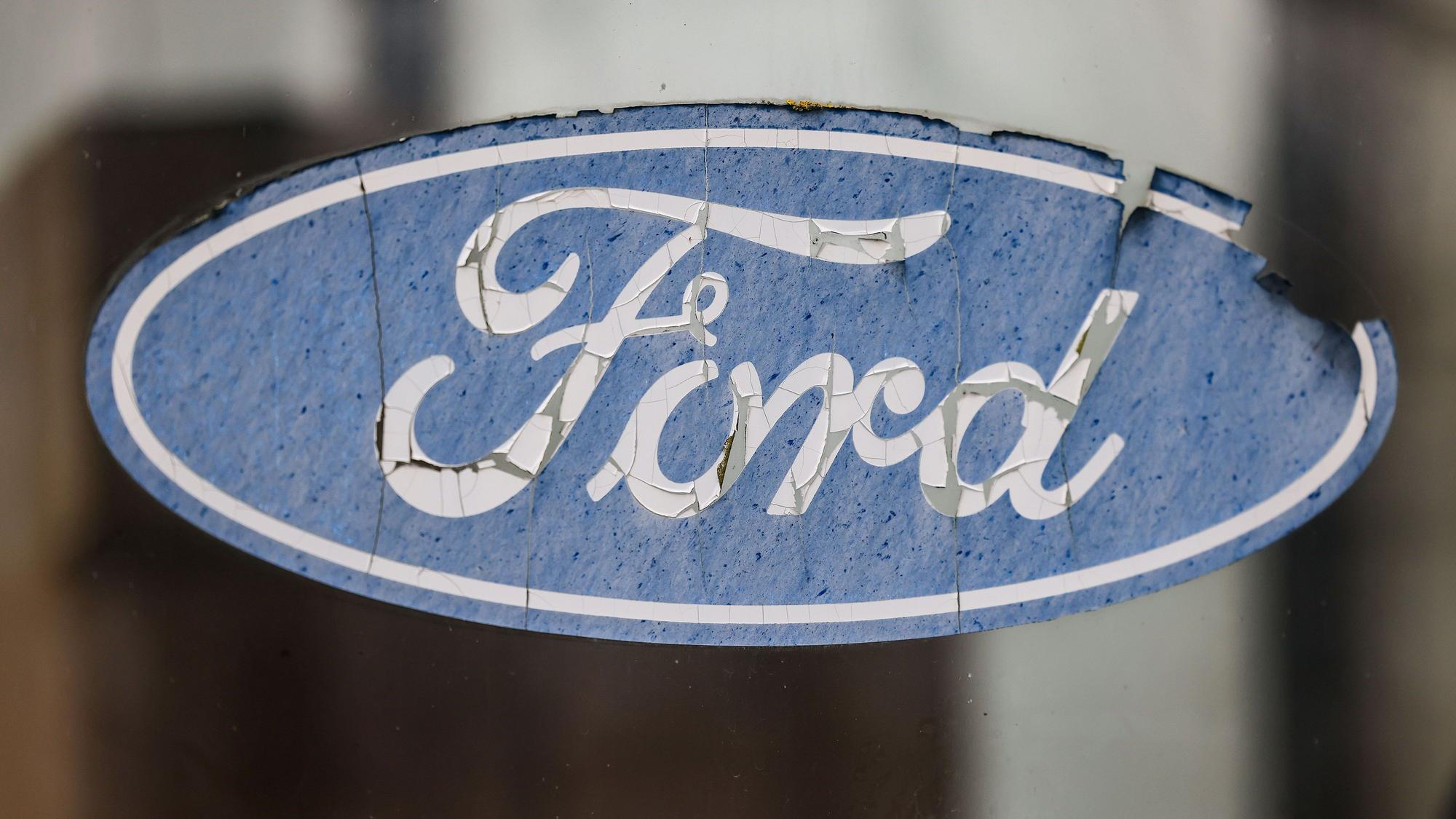 Ein abgeblättertes Logo von Ford hängt an einer Scheibe vor dem Werksgelände.