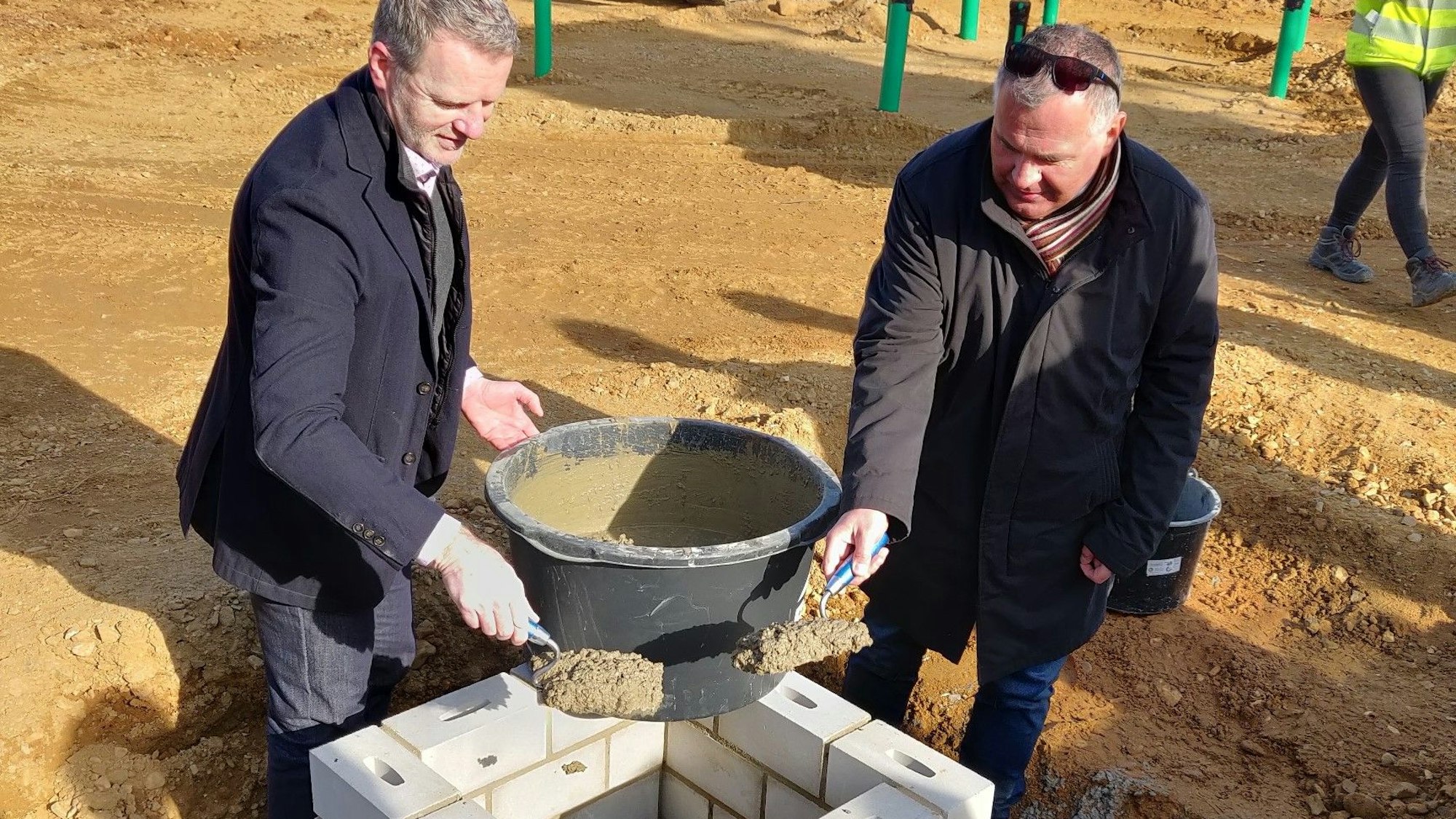 Bürgermeister Frank Steffes und Amtsleiter Stephan Bergmann füllen Mörtel mit Maurerkellen aus einem Eimer in den Grundsteinschacht.