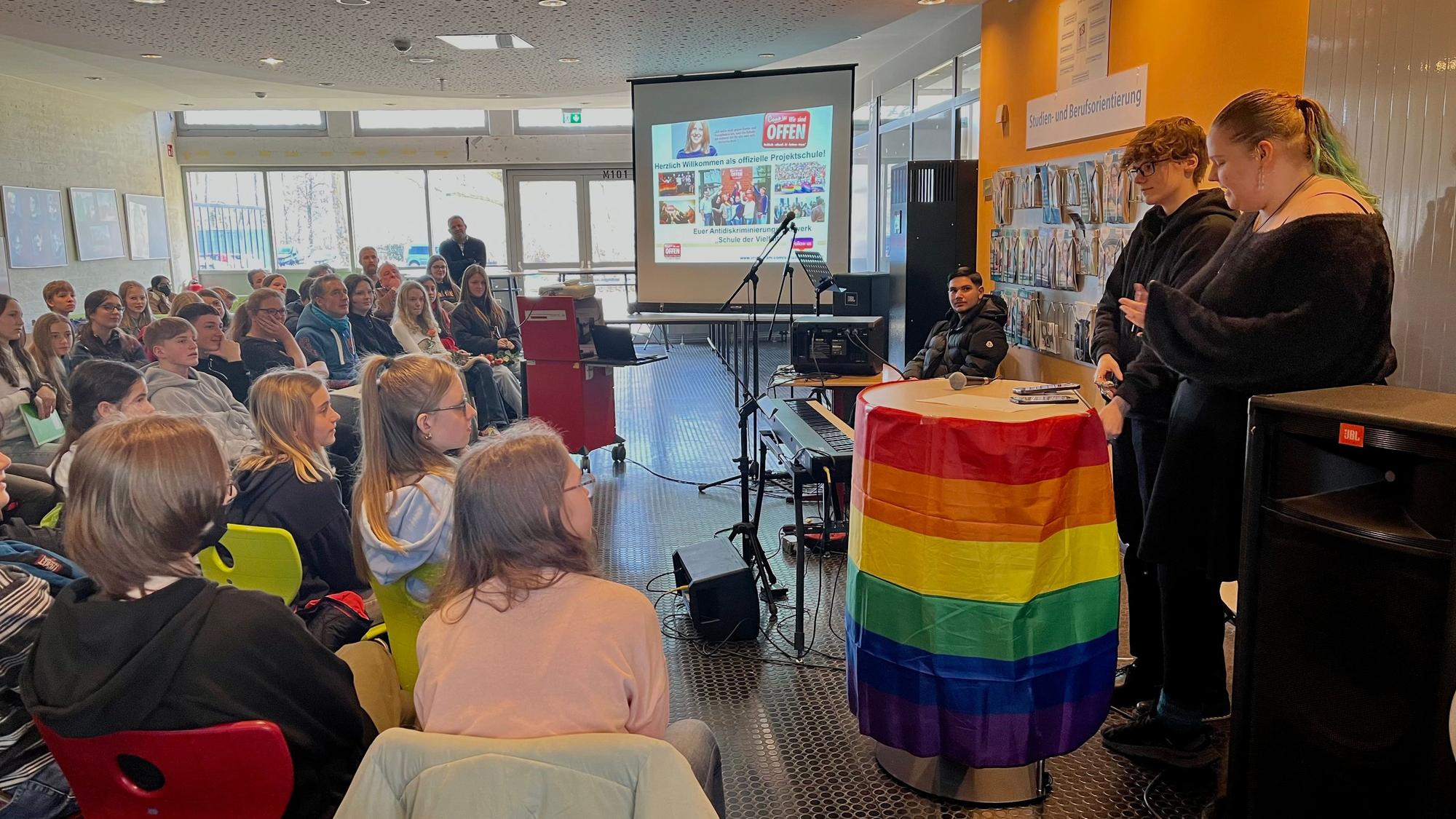 Blick in die Versammlung mit zwei Rednern an einem Pult in Regenbogenflagge