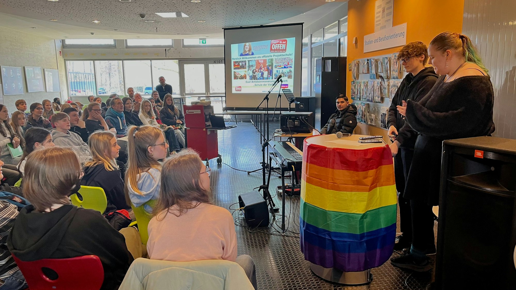 Blick in die Versammlung mit zwei Rednern an einem Pult in Regenbogenflagge