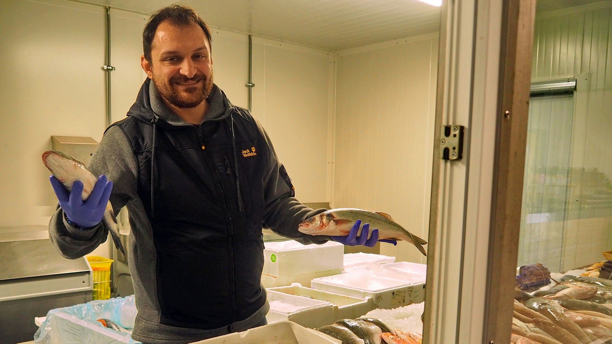 Der Verkäufer von Sakis Fisch und Feinkost hat zwei Fische in den Händen.