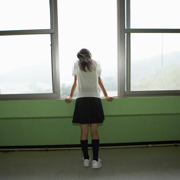 Ein Mädchen mit kurzem Rock und weißen Sneakers steht alleine an einem geöffneten Fenster.