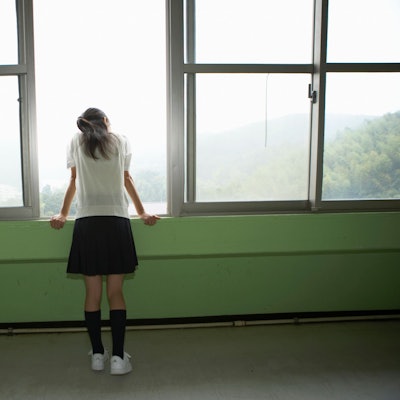 Ein Mädchen mit kurzem Rock und weißen Sneakers steht alleine an einem geöffneten Fenster.