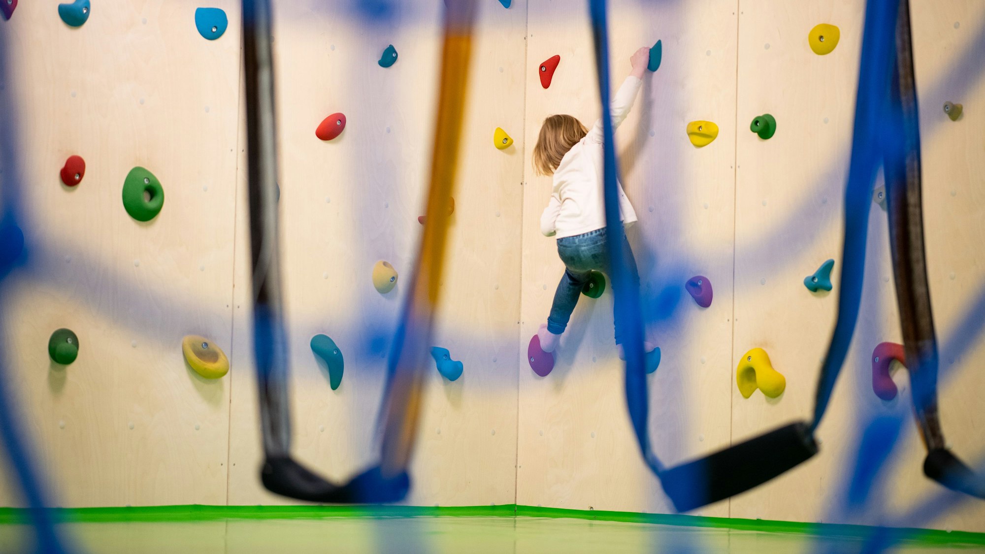 Ein Kind zieht sich an einer Kletterwand hoch. Davor sind Schlingen zu sehen.