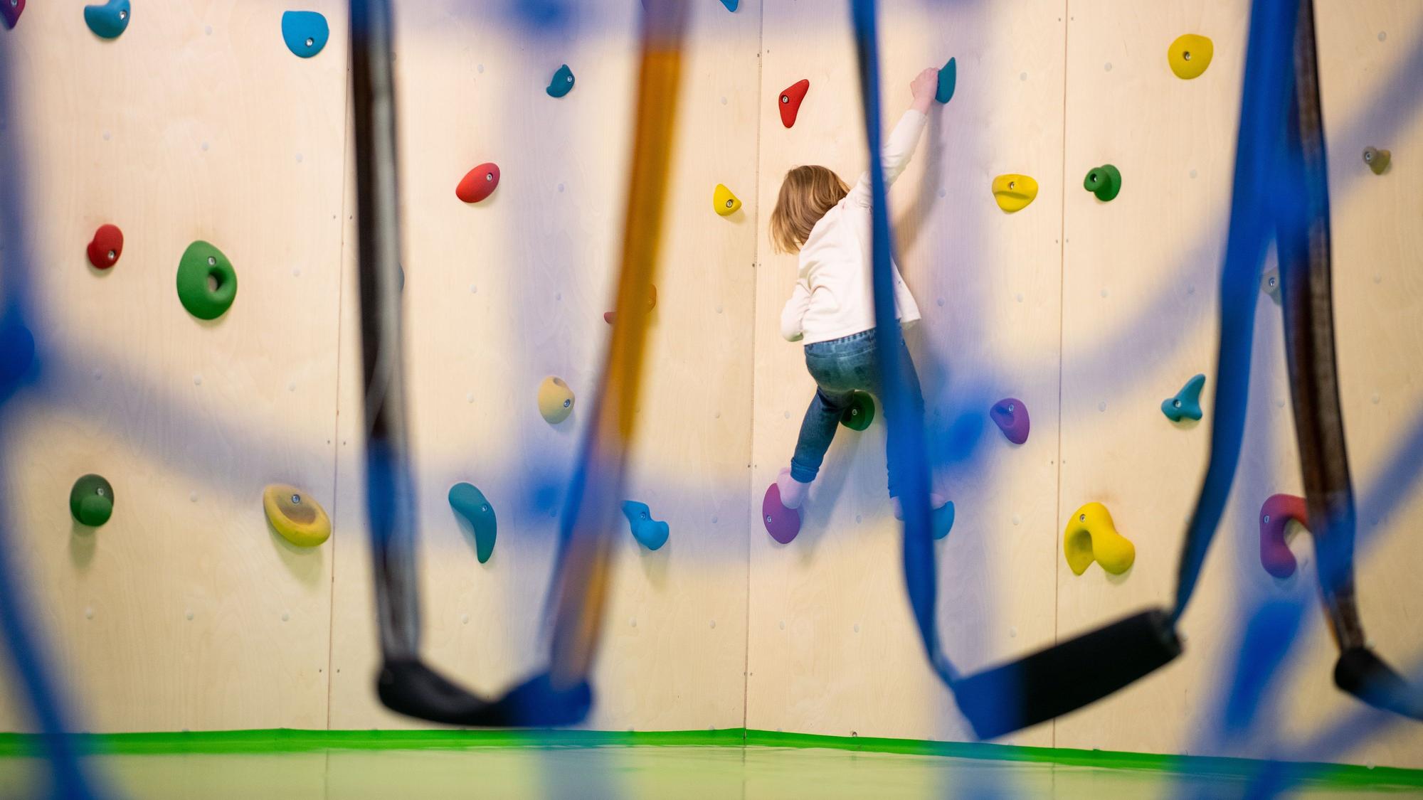 Ein Kind zieht sich an einer Kletterwand hoch. Davor sind Schlingen zu sehen.