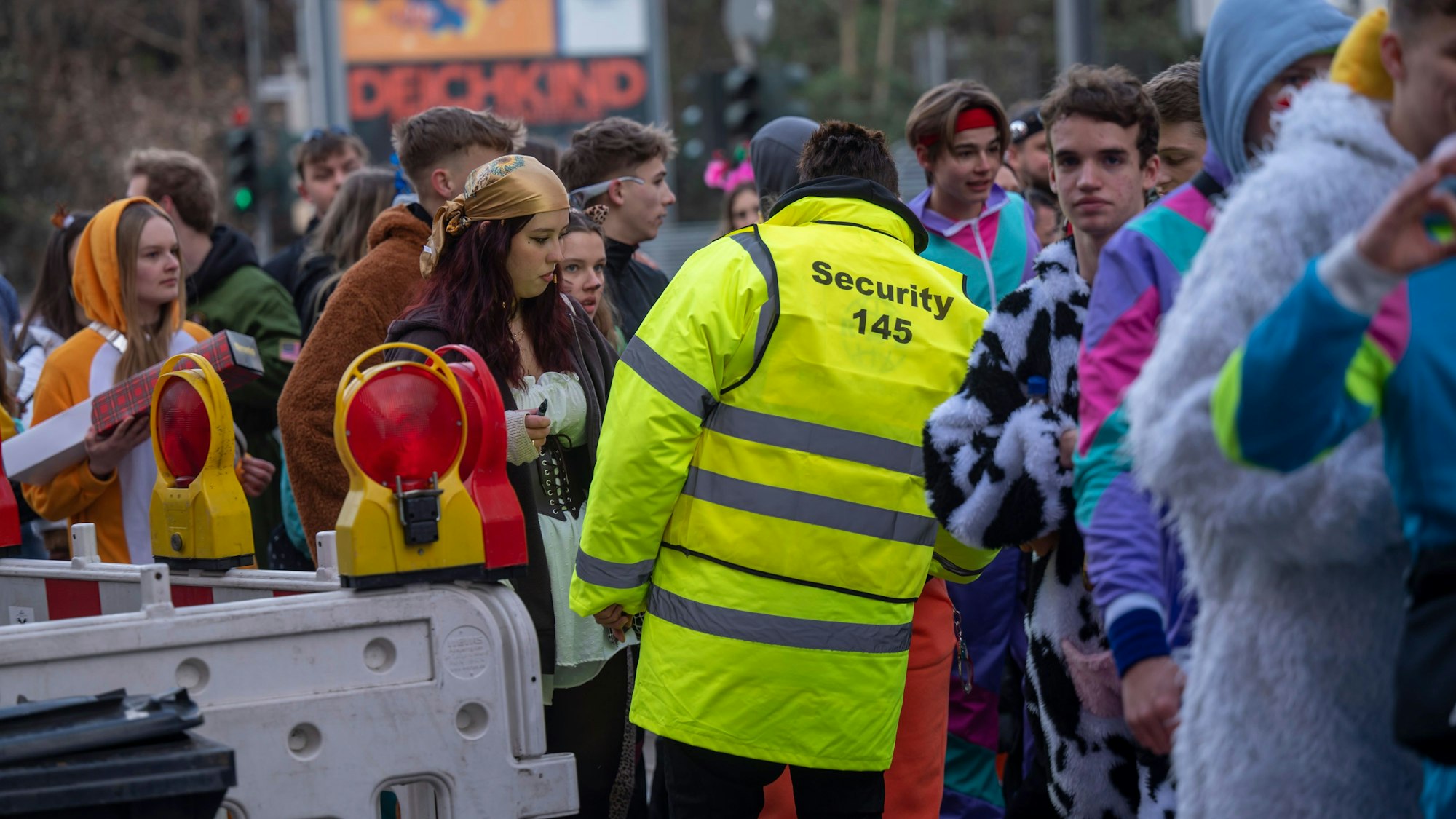 Ein Ordner eines privaten Sicherheitsdienstes und kostümierte Menschen an einer Absperrung im Kwartier Latäng an Weiberfastnacht 2023