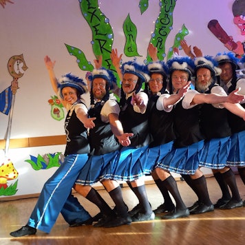 Eine Tanzgruppe steht auf einer Bühne in einer Reihe hintereinander und schaut mit ausgebreiteten Armen in Richtung des Publikums.