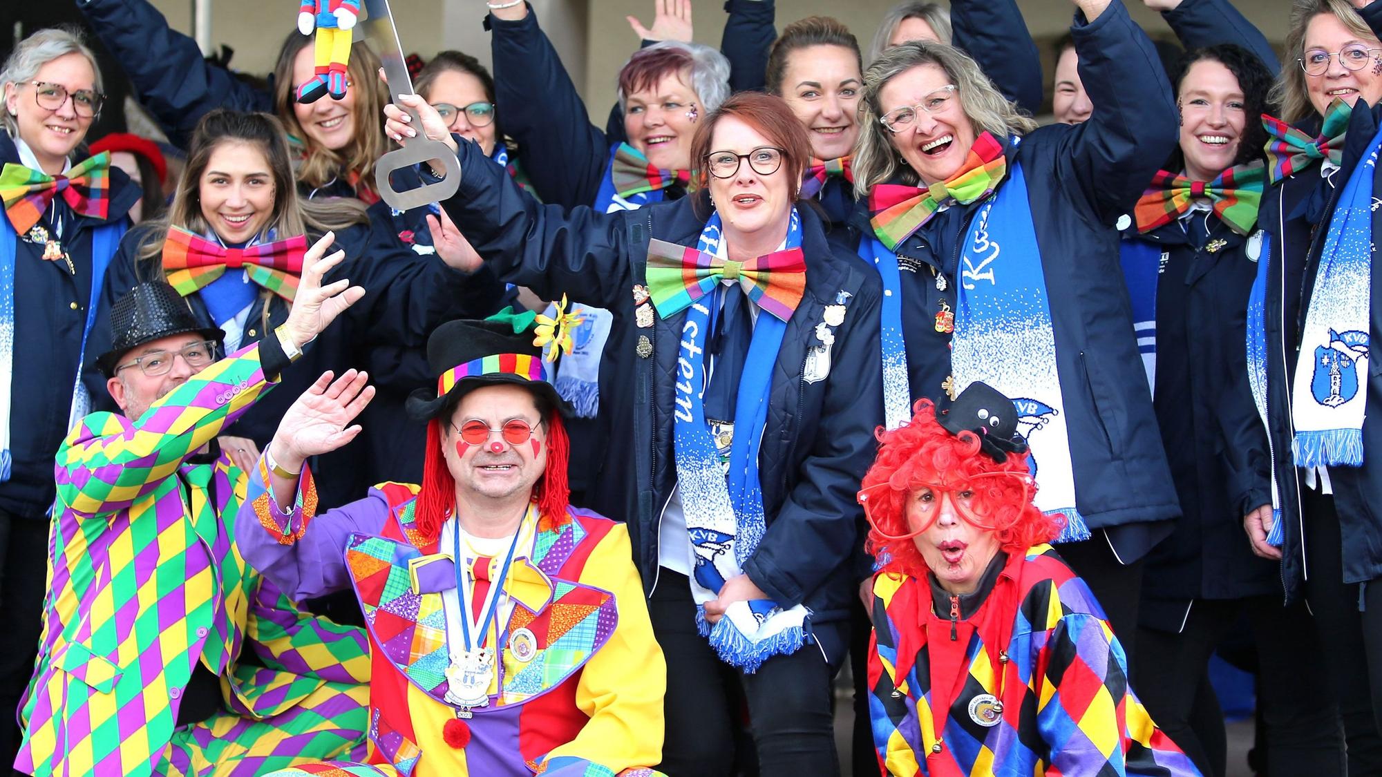 Ein als bunter Clown verkleideter Bürgermeister sitzt inmitten Feiernder und ebenfalls verkleideter Frauen.