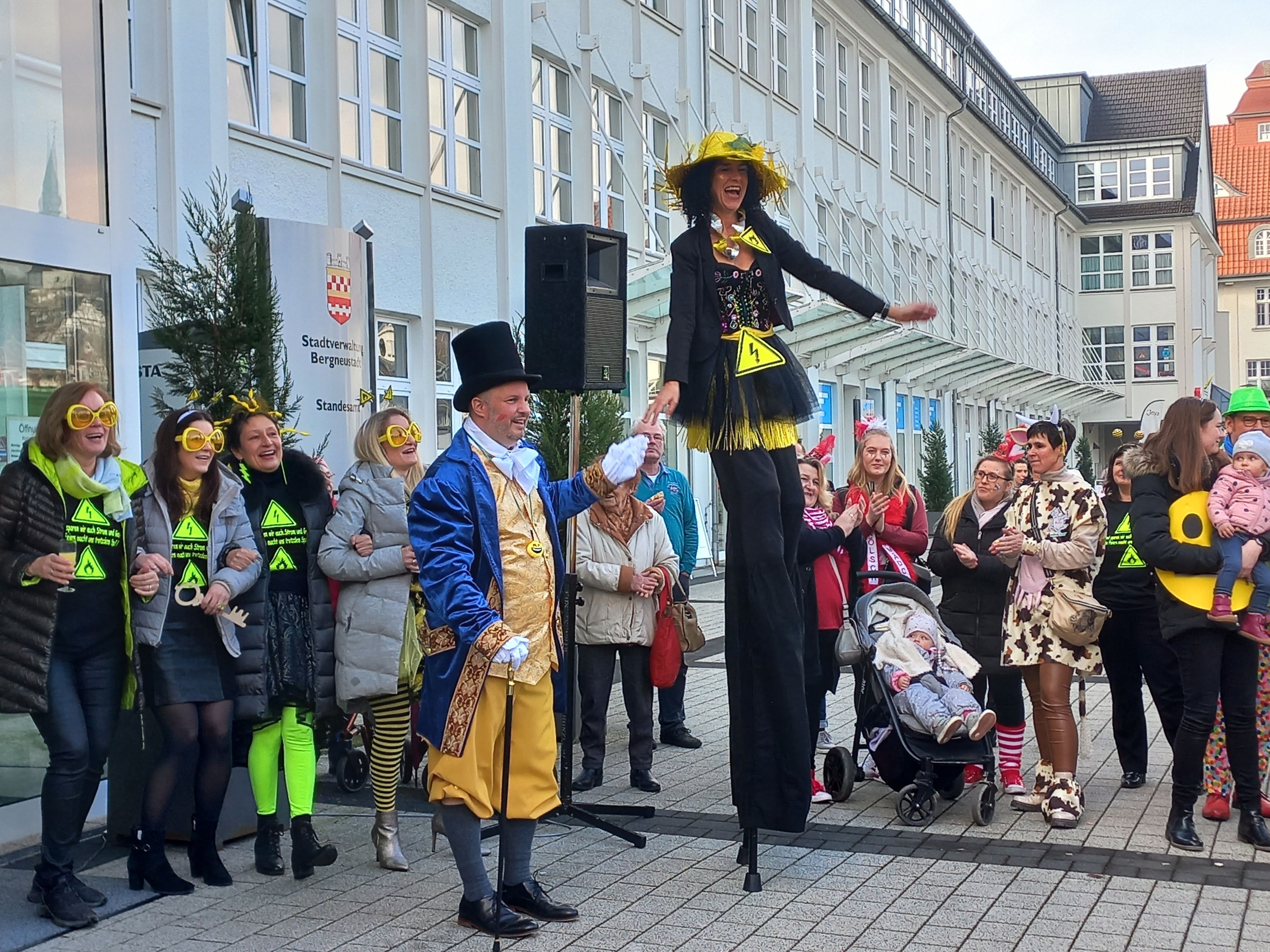 Eine verkleidete Frau steht auf langen Stelzen und überragt weitere verkleidete Menschen. Ein Mann mit Zylinder hält sie an der Hand.