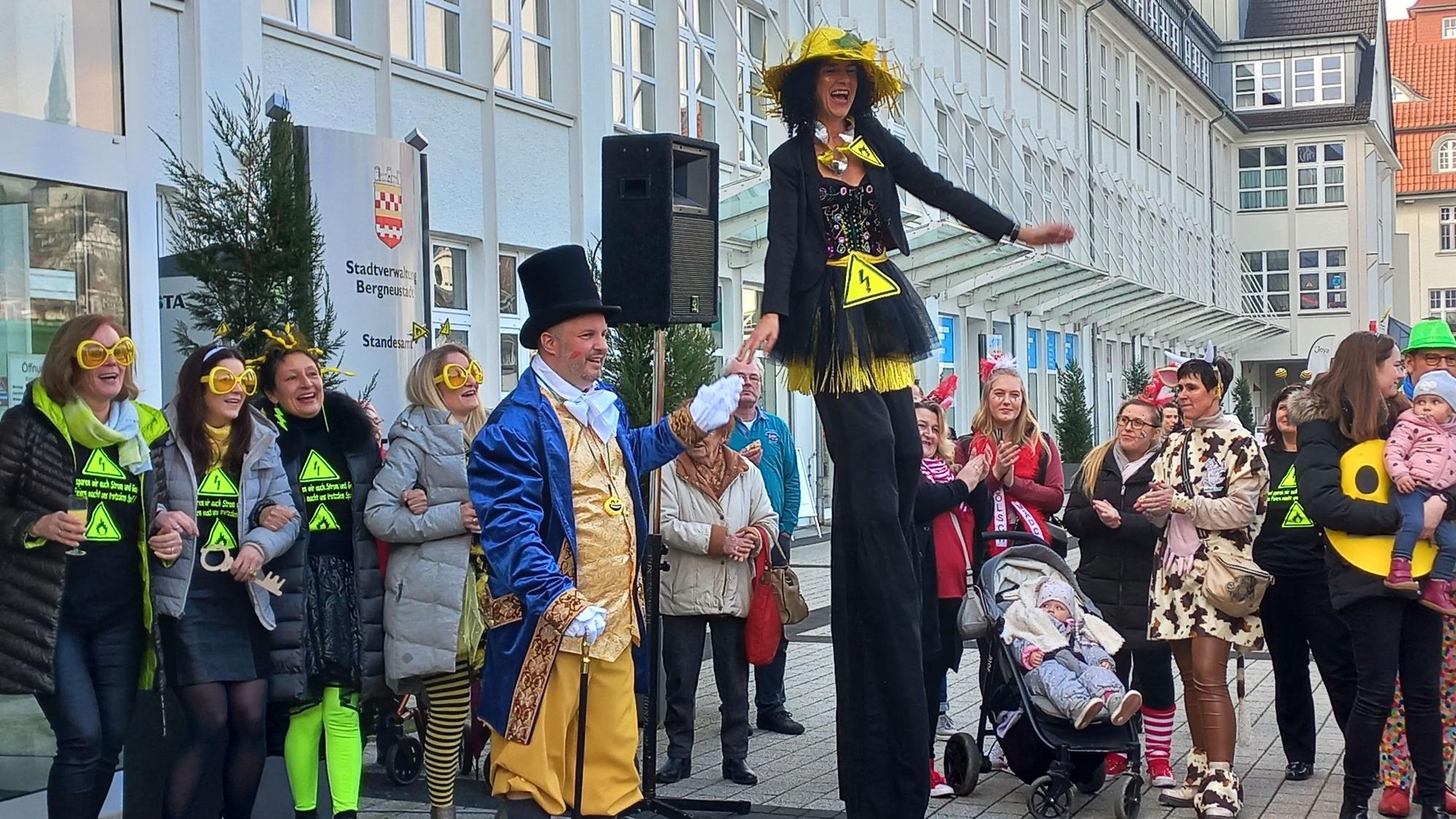 Eine verkleidete Frau steht auf langen Stelzen und überragt weitere verkleidete Menschen. Ein Mann mit Zylinder hält sie an der Hand.