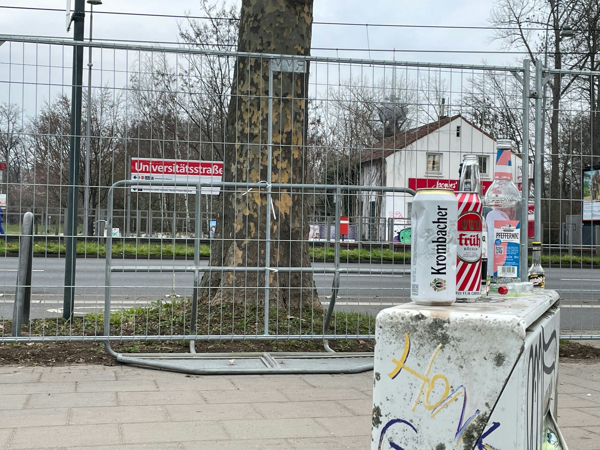 Bierdosen stehen auf einem Elektronikkasten am Straßenrand. Dahinter ist die Bahnhaltestelle „Universitätsstraße“, die eingezäunt ist.